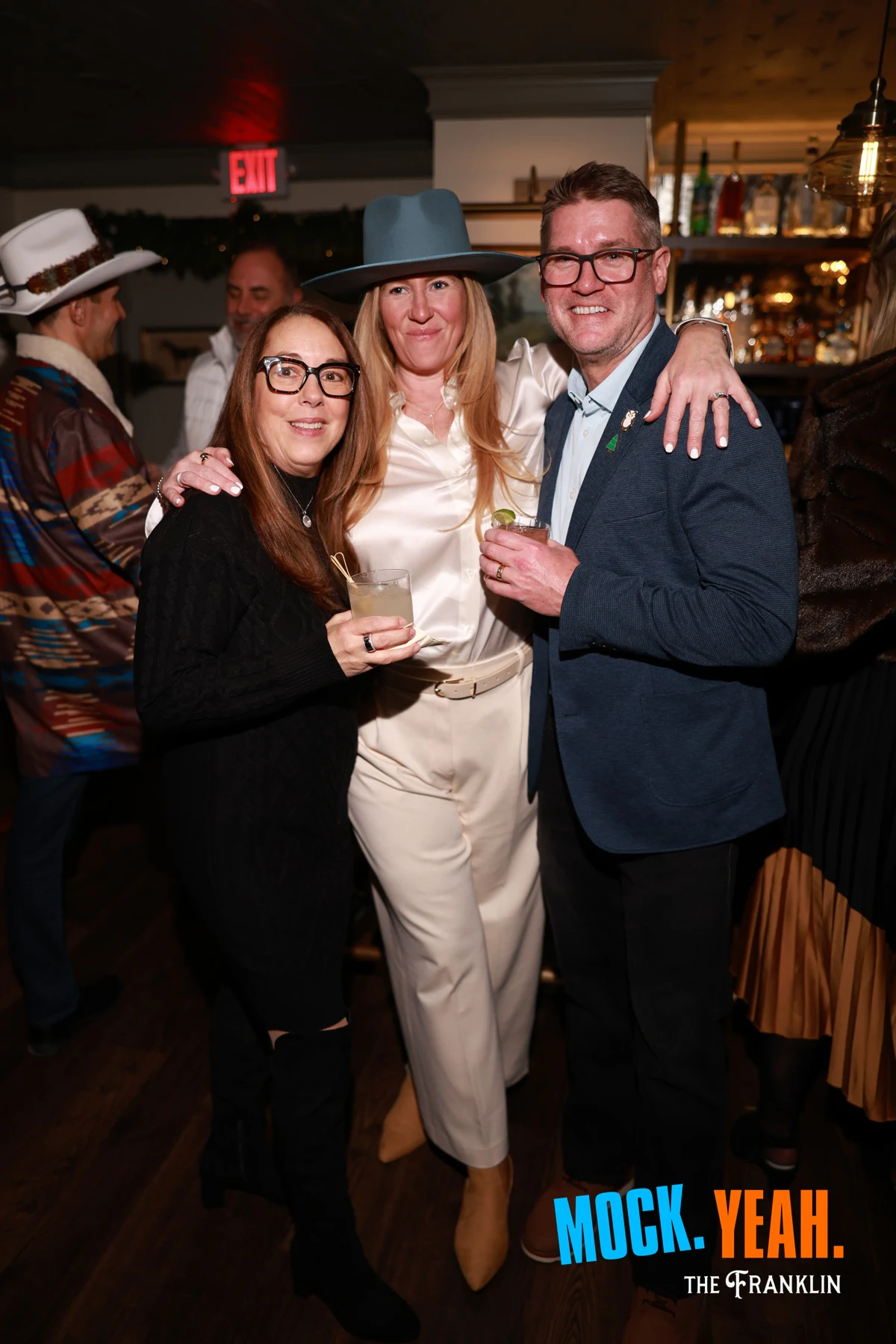 Three guests with drinks, the one in the middle in a wide-brim hat at the holiday party at The Franklin