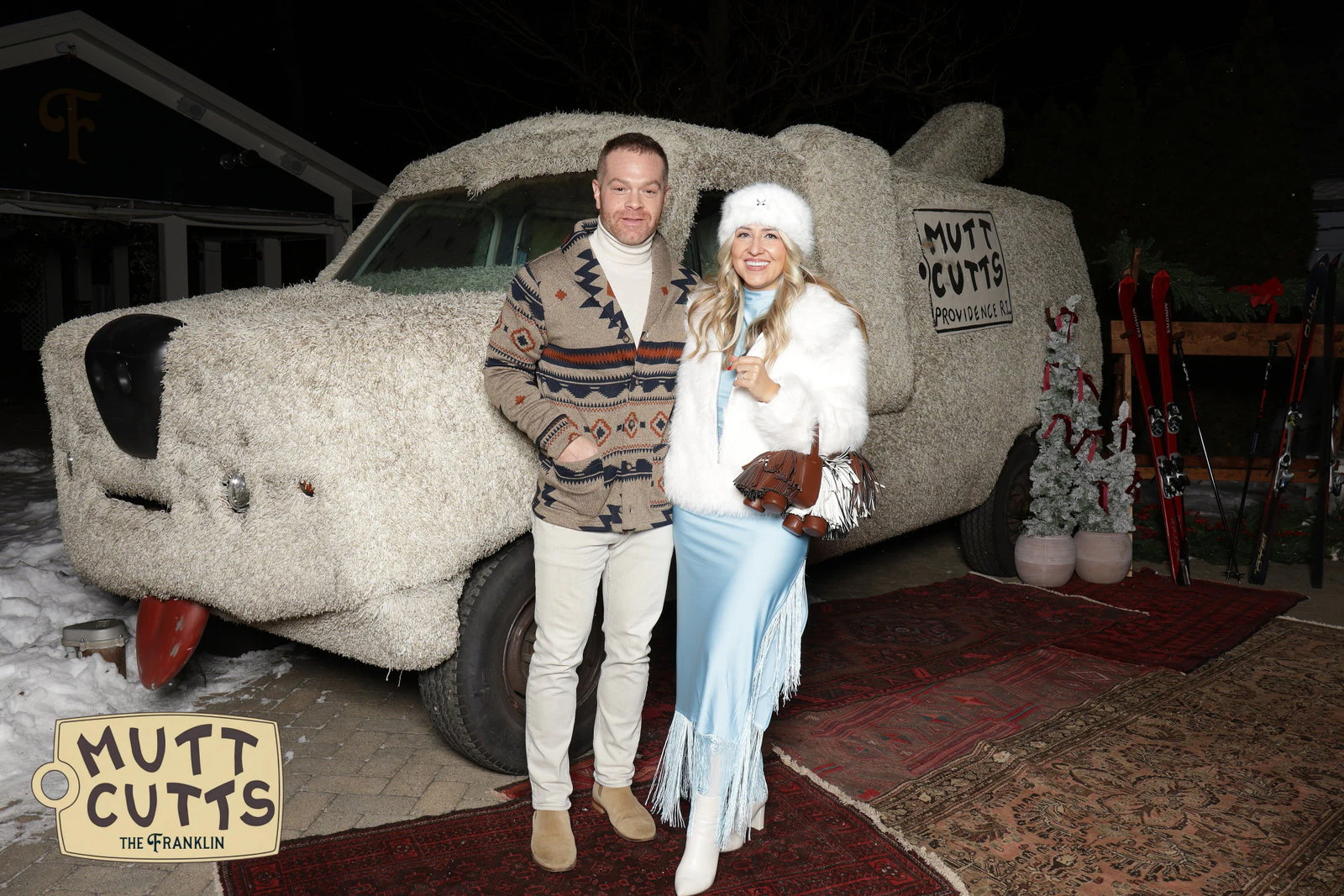 A man in a patterned cardigan and a woman in a white fur hat and jacket and a pale blue fringed skirt in front of the shaggy Mutt Cutts van at The Franklin Holiday Party