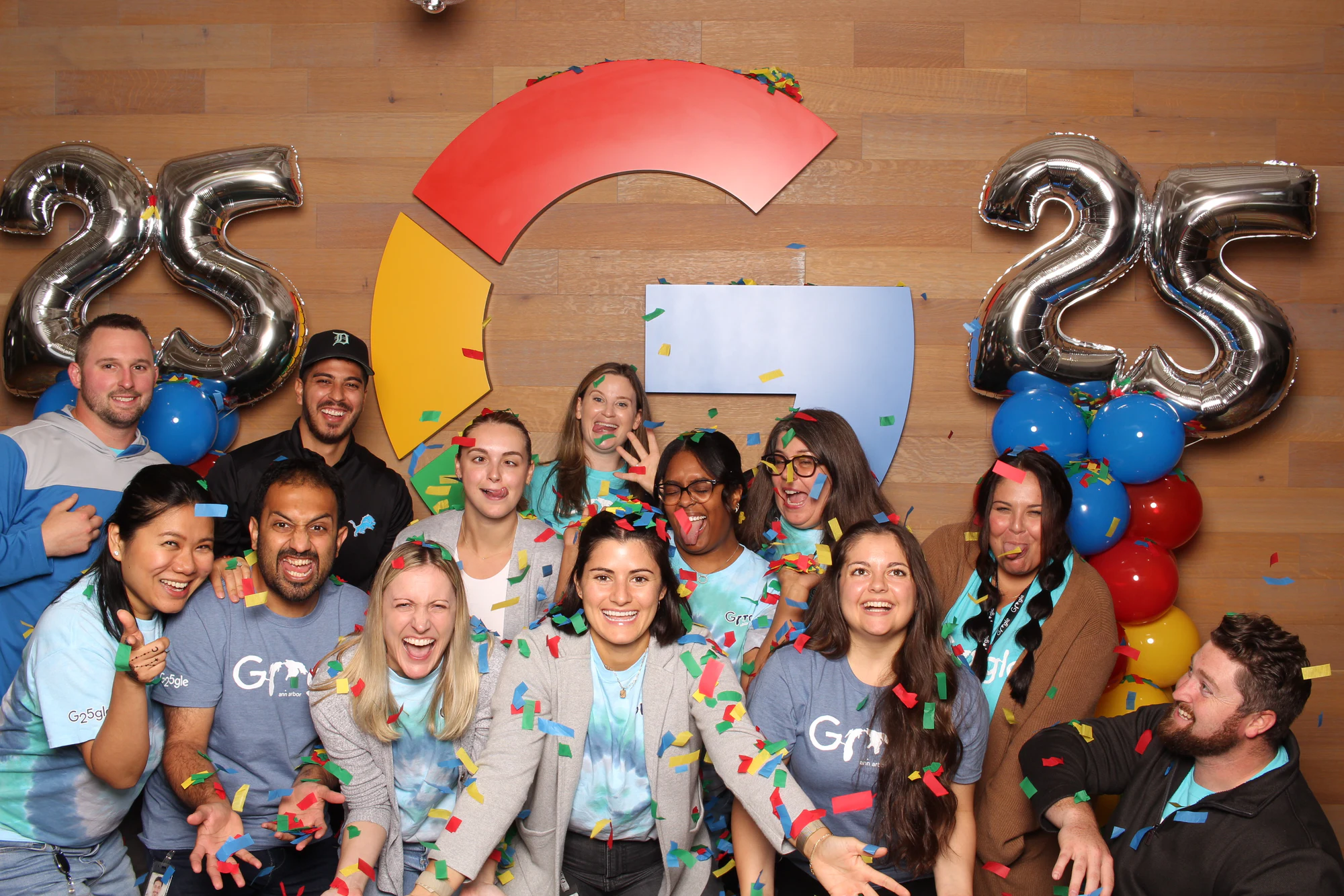 A big team packed in front of the Google logo and silver 25 balloons, laughing at Google’s 25th Birthday