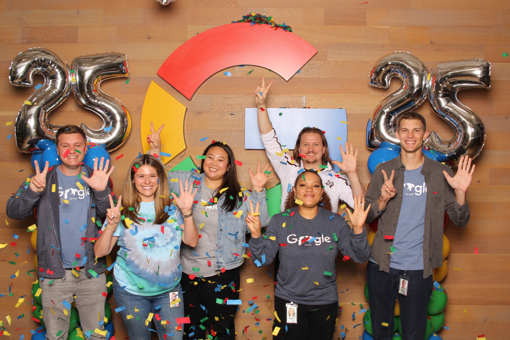 Nine coworkers in Google shirts and tie-dye waving under falling confetti at Google’s 25th Birthday
