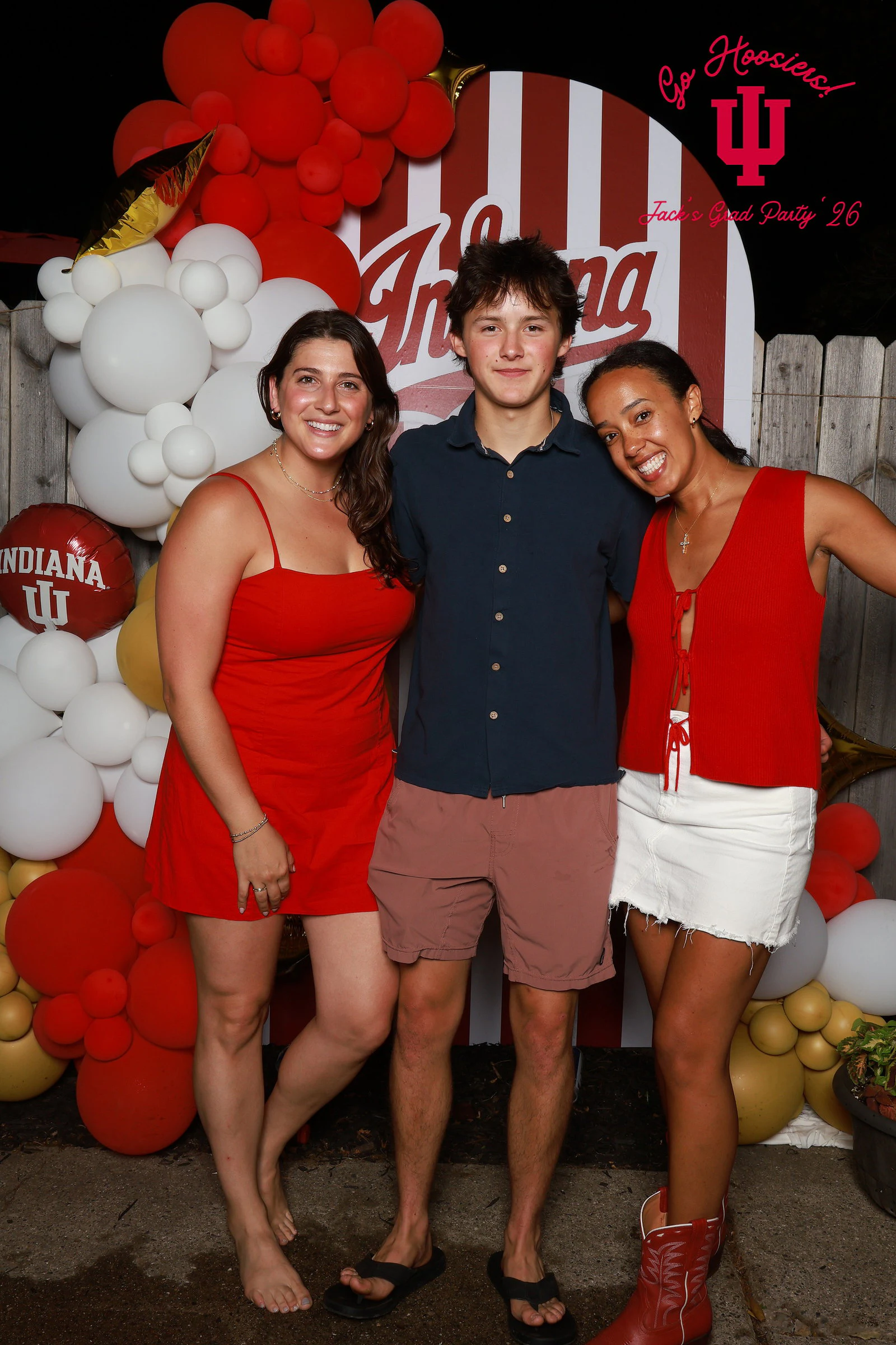 A woman in a red dress, a young man in a navy shirt and a woman in a red top and white skirt in front of a red and white striped arch and balloon garland at a private party