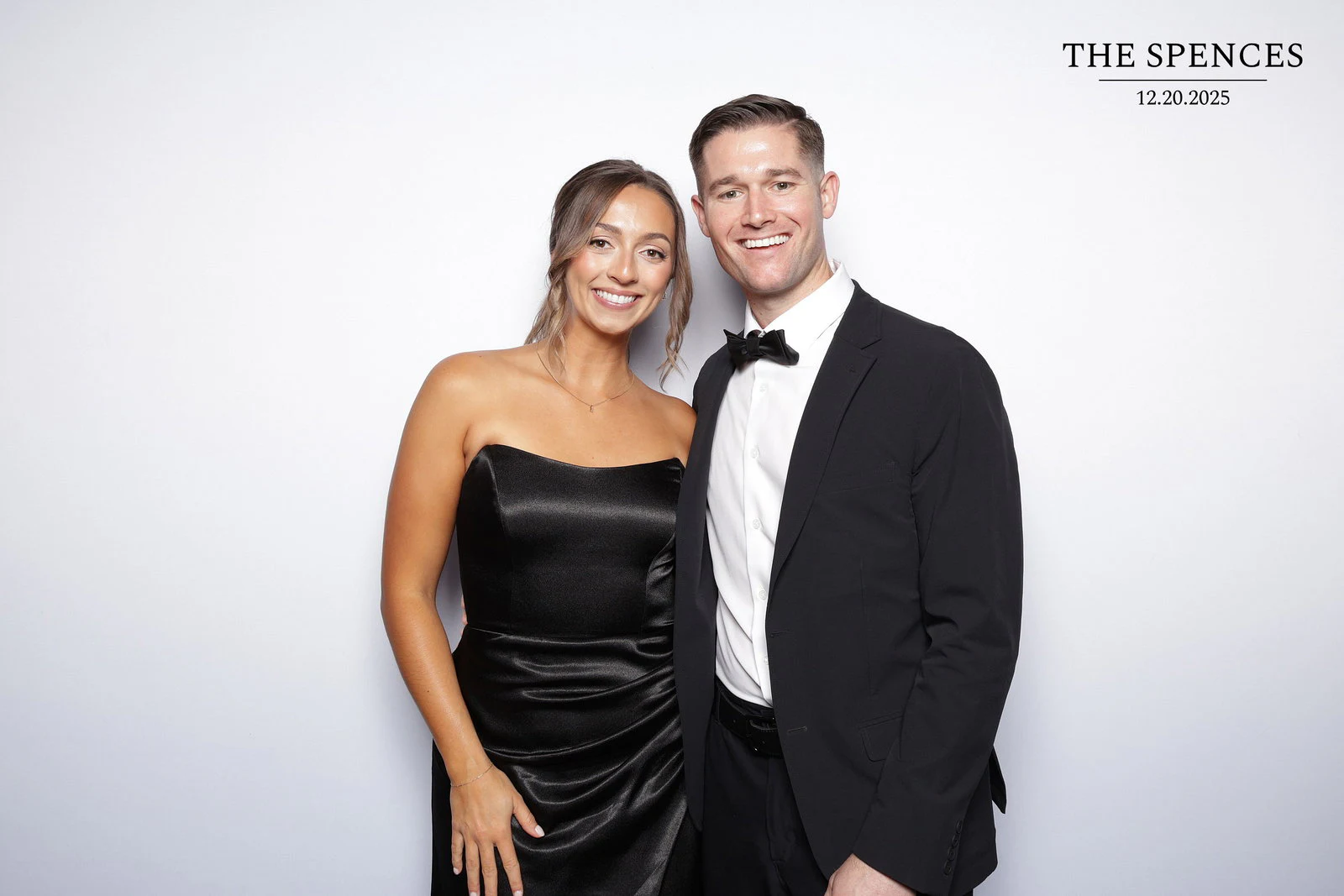 A woman in a black satin strapless dress and a man in a tuxedo smiling in front of the pure white backdrop at the Jessica + Rory wedding