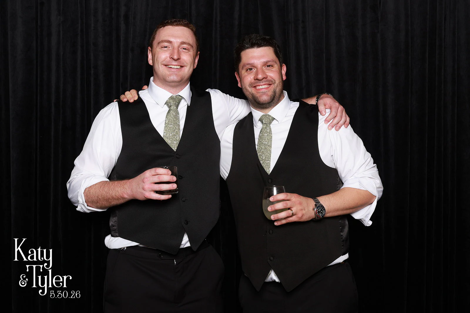 Two men in matching black waistcoats and sage ties, arms over each other’s shoulders, drinks in hand at Katy + Tyler