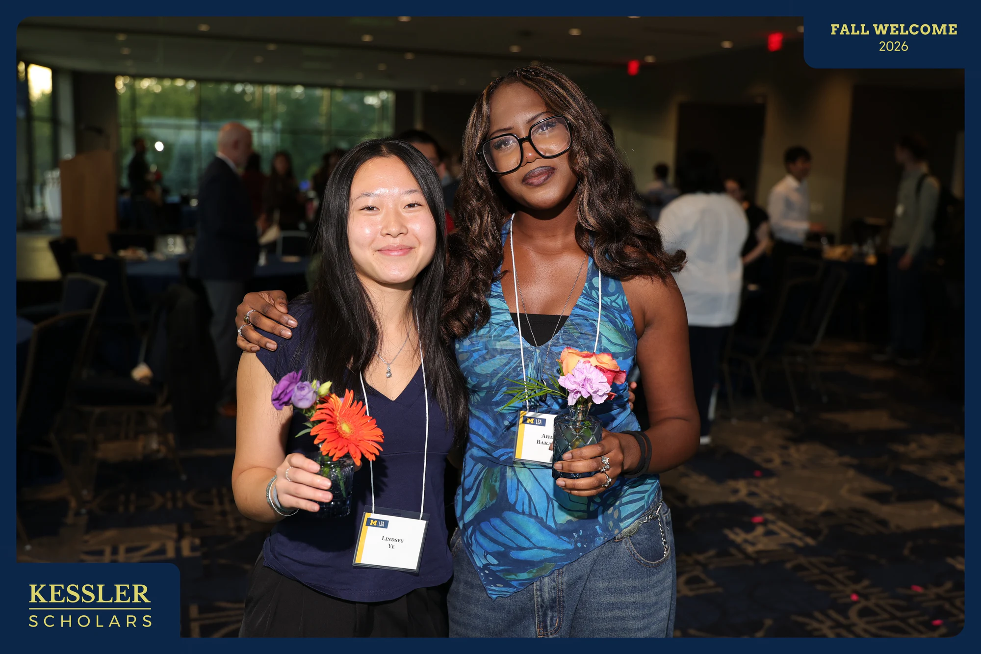 Two students holding small vases of flowers, one with an arm around the other at Kessler Fall Welcome Dinner