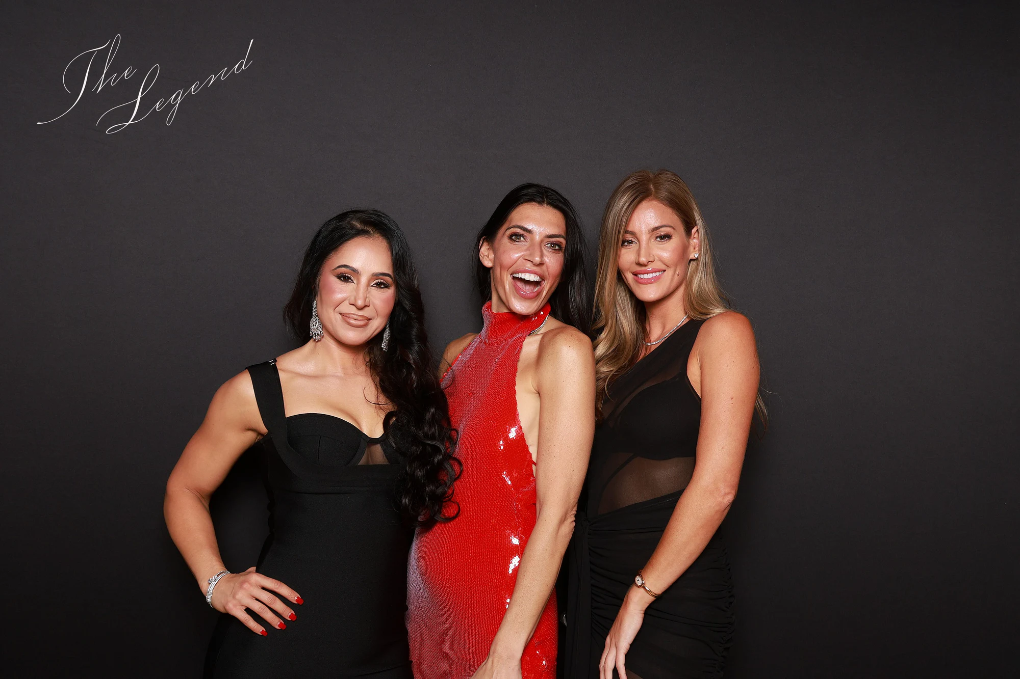 Three women dressed up, a red sequin dress in the middle, one mid-laugh, against the plain black backdrop at a birthday party