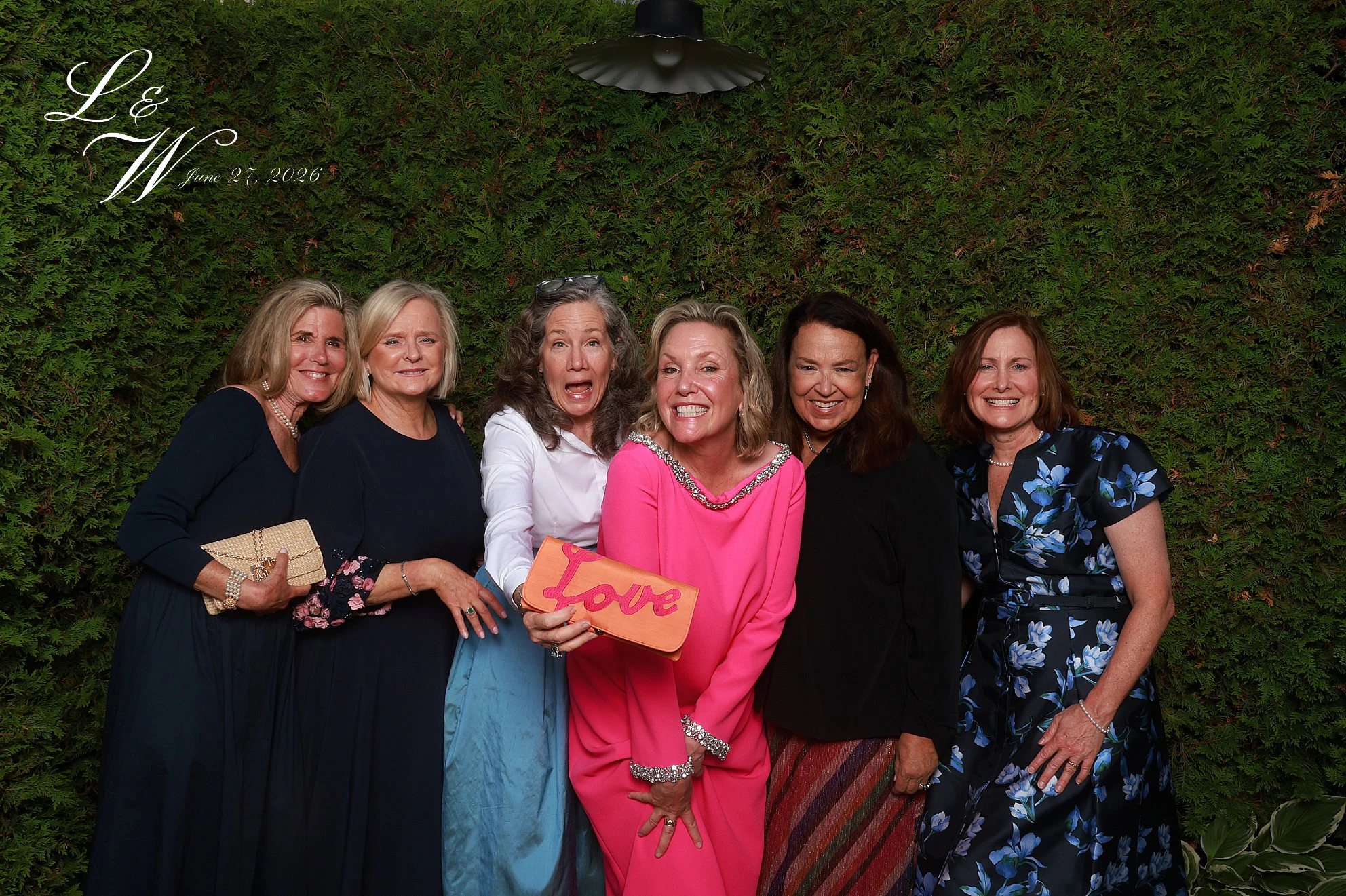 Six women in a row in front of the hedge, one in a hot pink dress holding a clutch that says Love at Lindsay + Will
