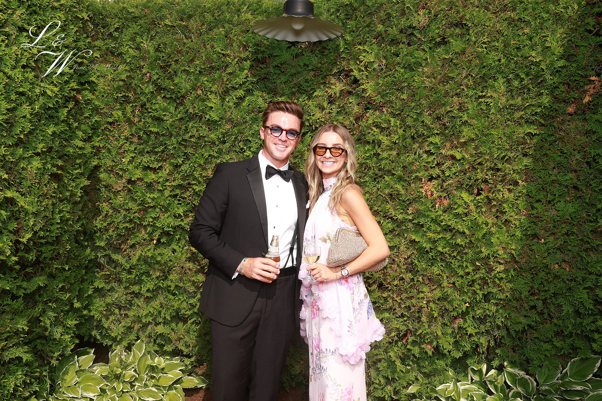 A man in a tuxedo and sunglasses with a woman in a pink ruffled dress, both holding drinks in front of the hedge at Lindsay + Will