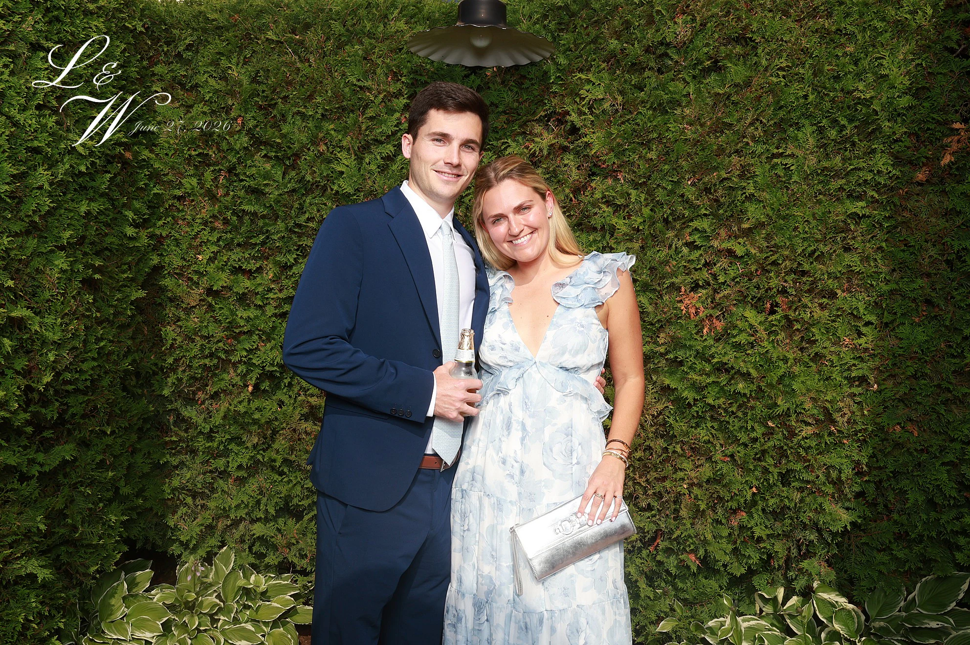 A man in a navy suit with his arm around a woman in a blue floral dress in front of the hedge at Lindsay + Will