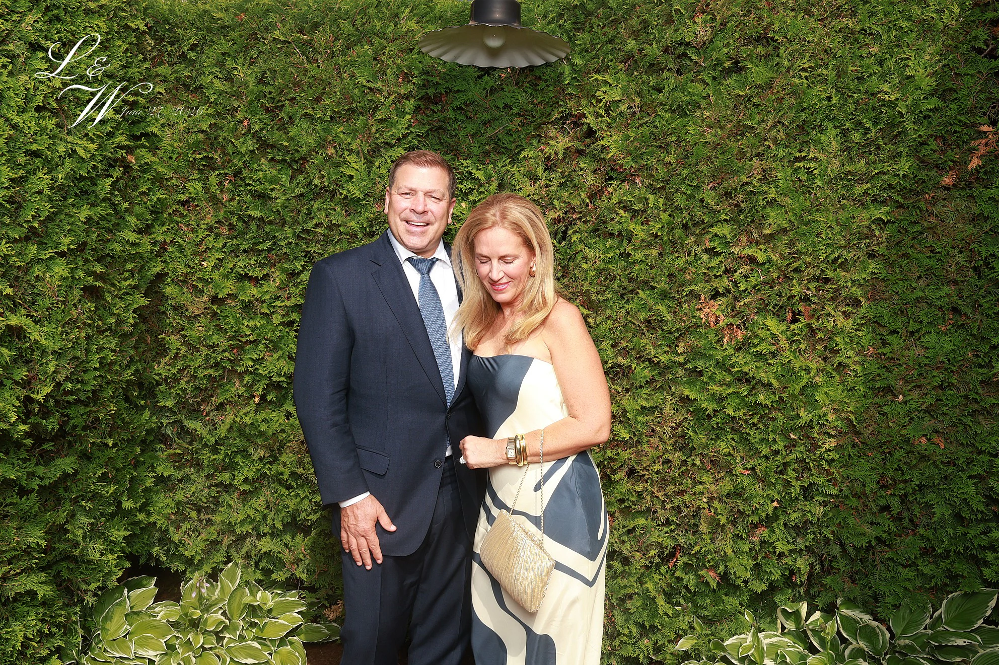 A man in a navy suit and a woman in a navy and cream print dress standing close in front of the hedge at Lindsay + Will