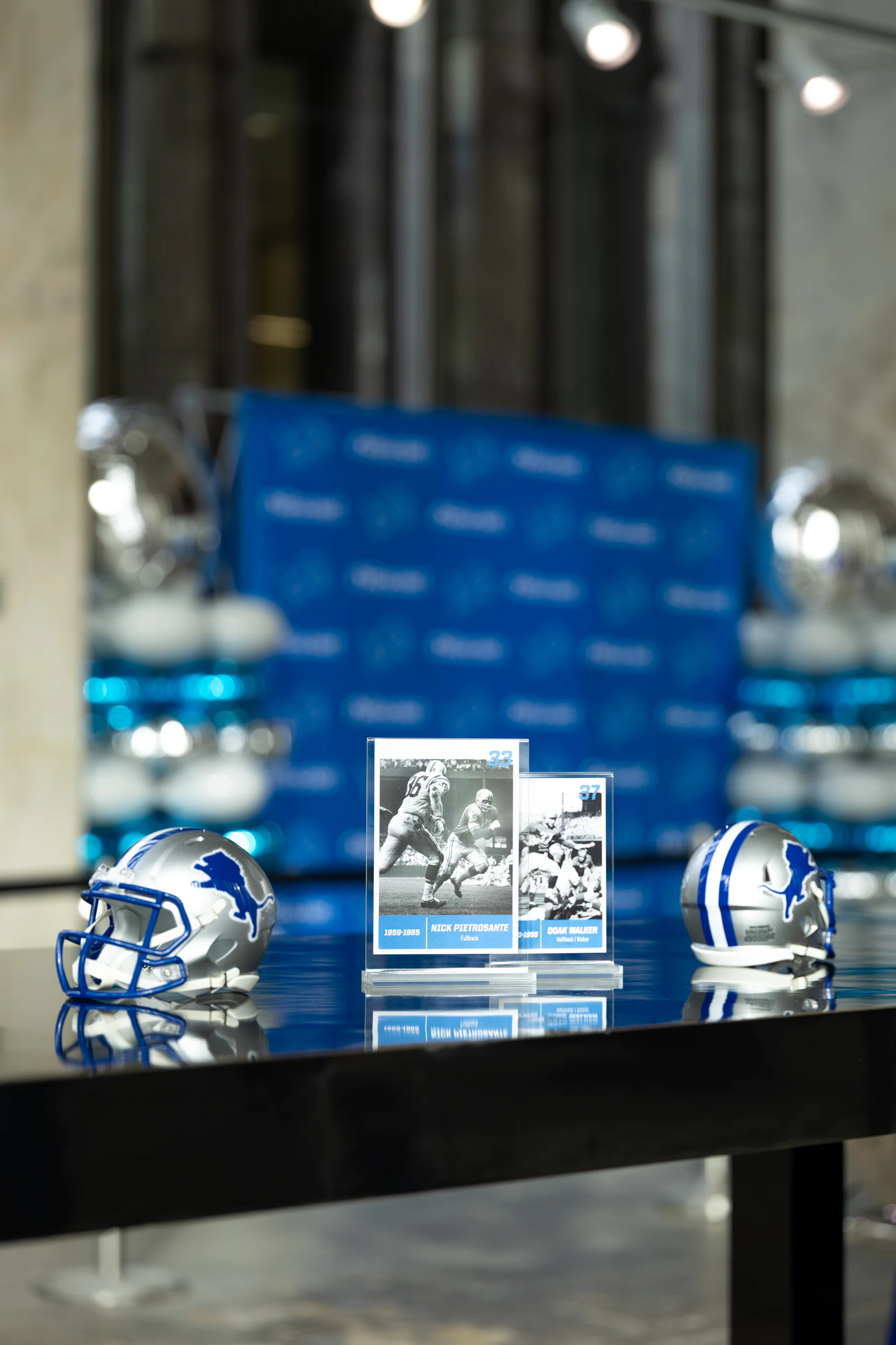 Two silver Lions helmets on a black table flanking a framed card of Nick Pietrosante and Doak Walker, a blue Rocket step-and-repeat behind