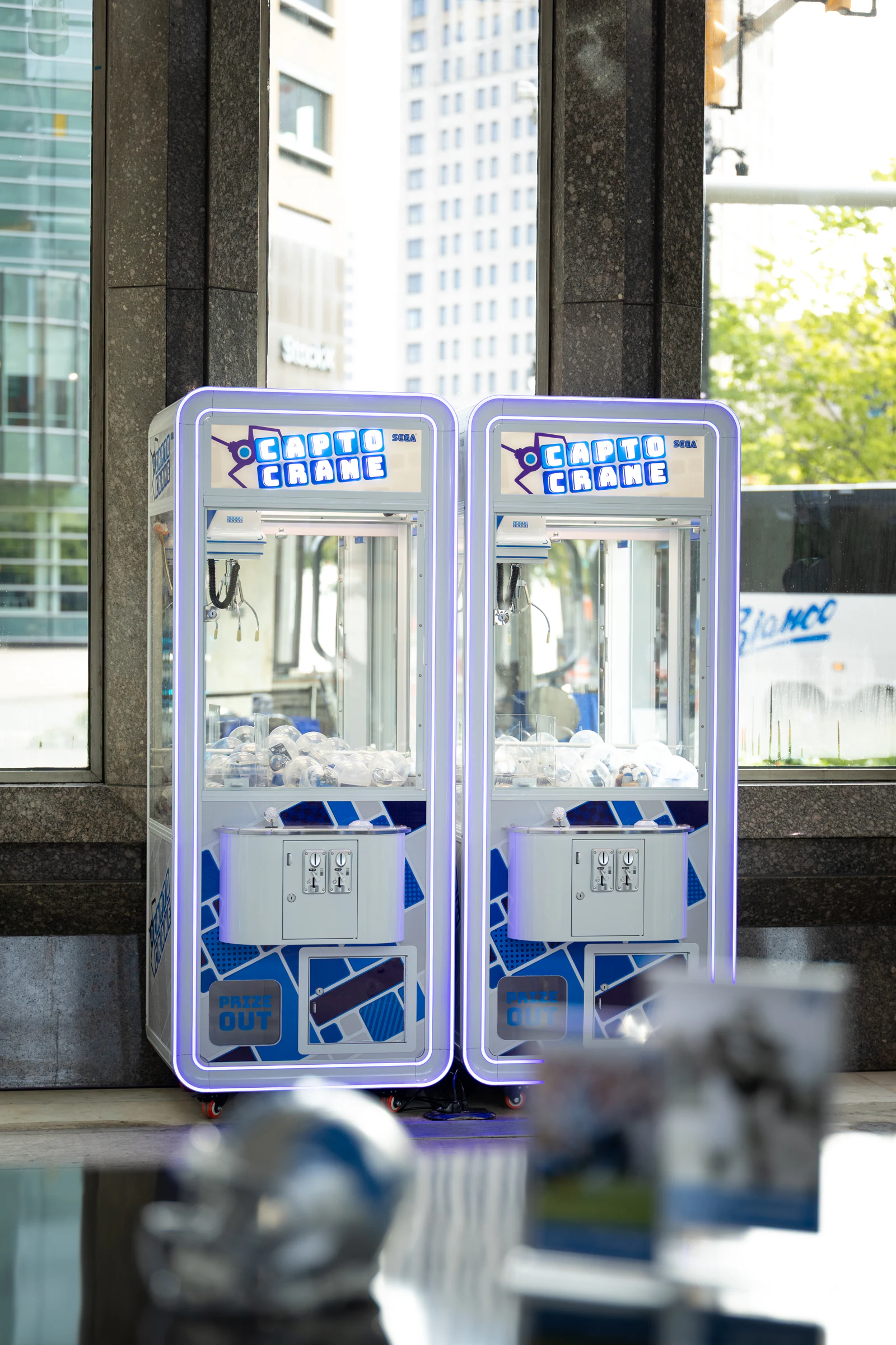 Two claw machines glowing purple in the lobby window, stocked with prizes, downtown Detroit outside