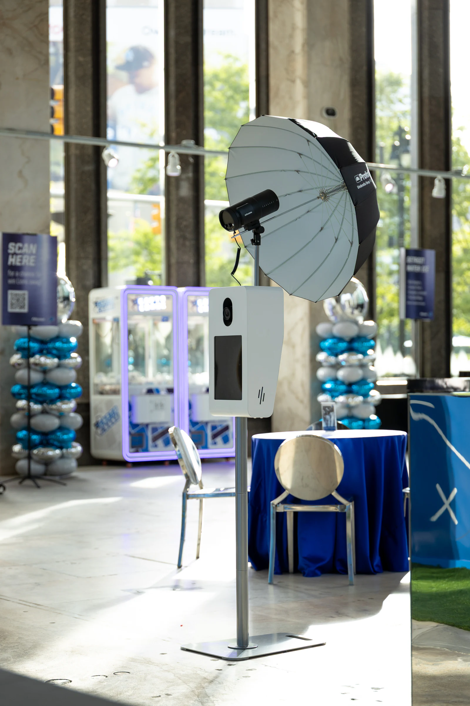 The Portrait Booth with its silver umbrella beside the claw machines, a blue-draped table and clear chairs waiting for guests