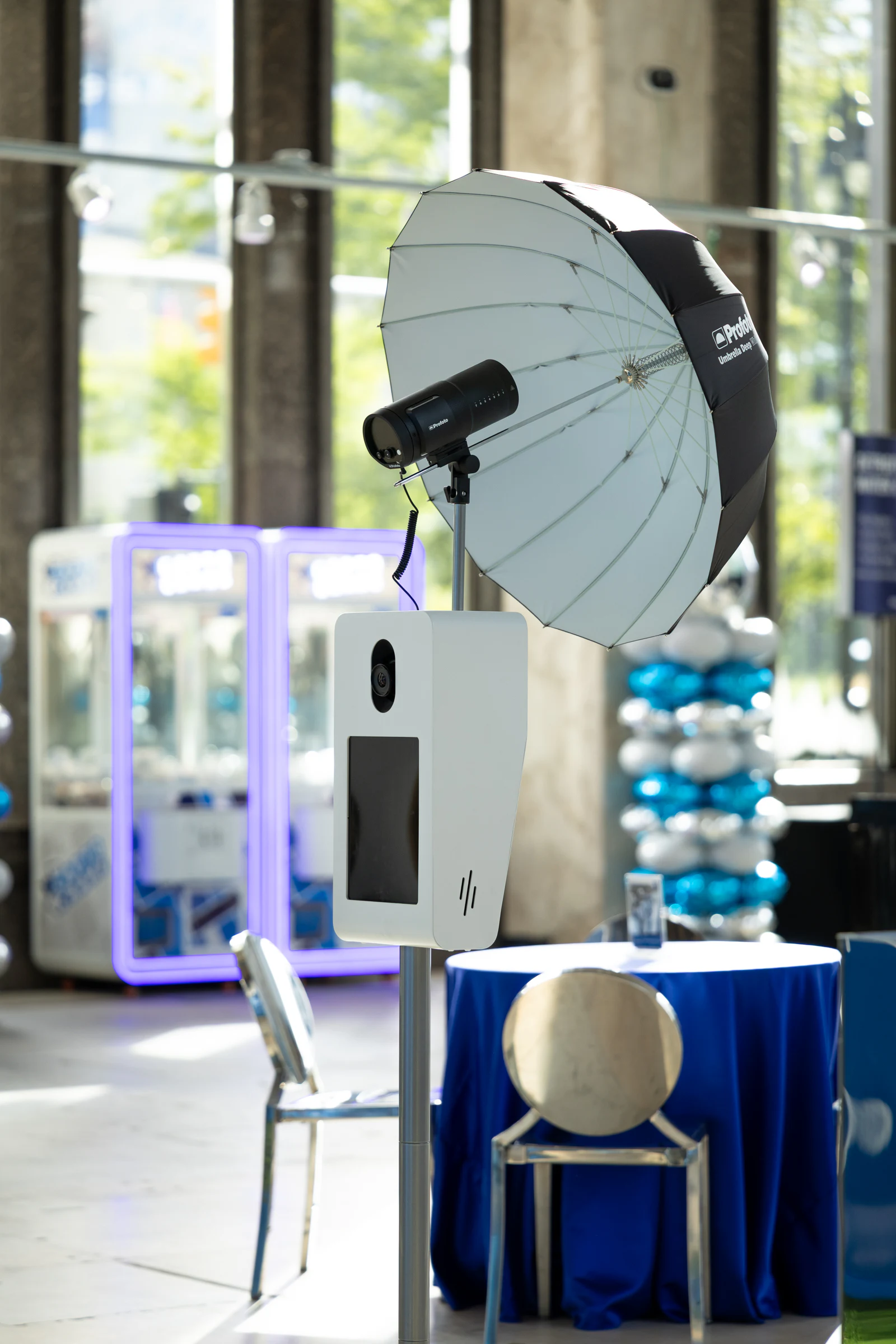 The booth's camera and umbrella lit by daylight through the lobby's tall windows, balloon towers and a blue table behind