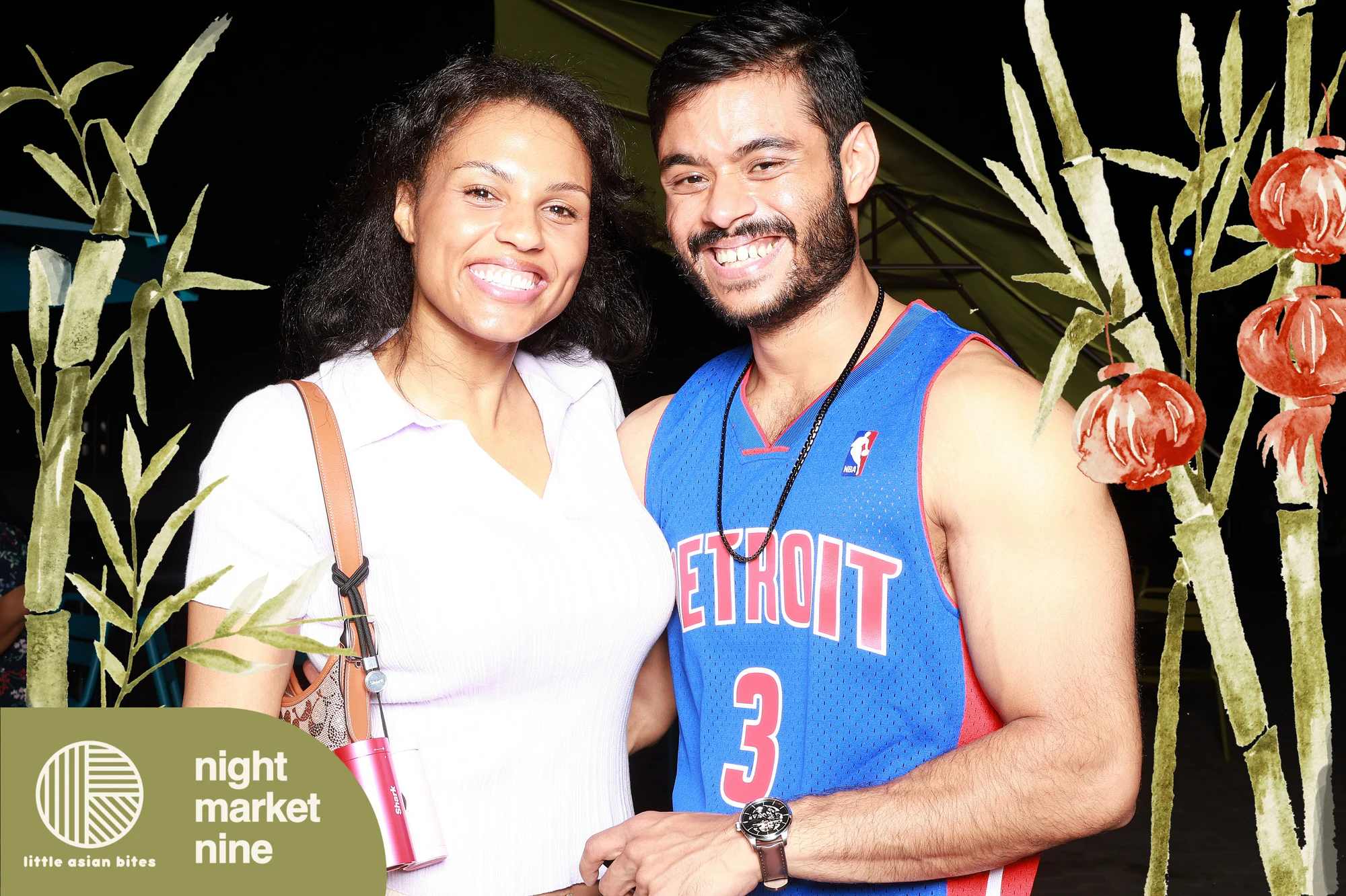 Couple in a Pistons jersey and white knit, both smiling straight down the lens at Little Asian Bites Night Market