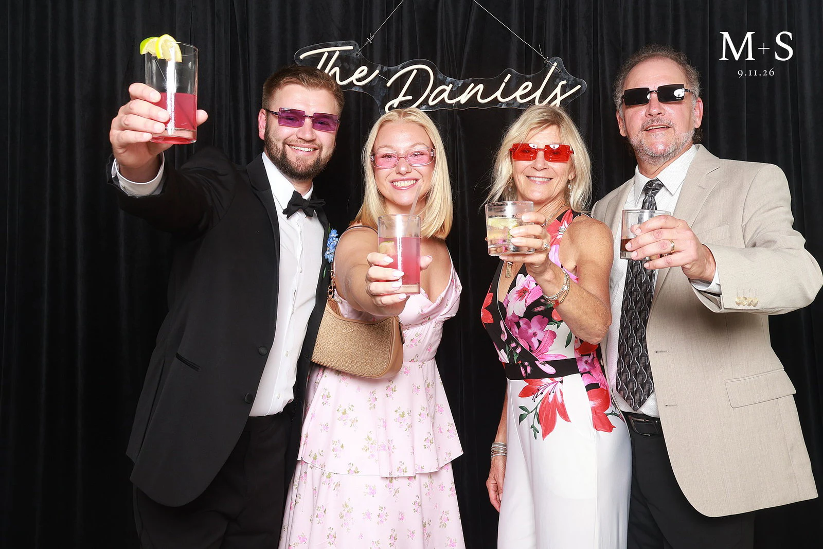 Four guests in coloured sunglasses holding their drinks out toward the camera, one raised high at Mariah + Seth