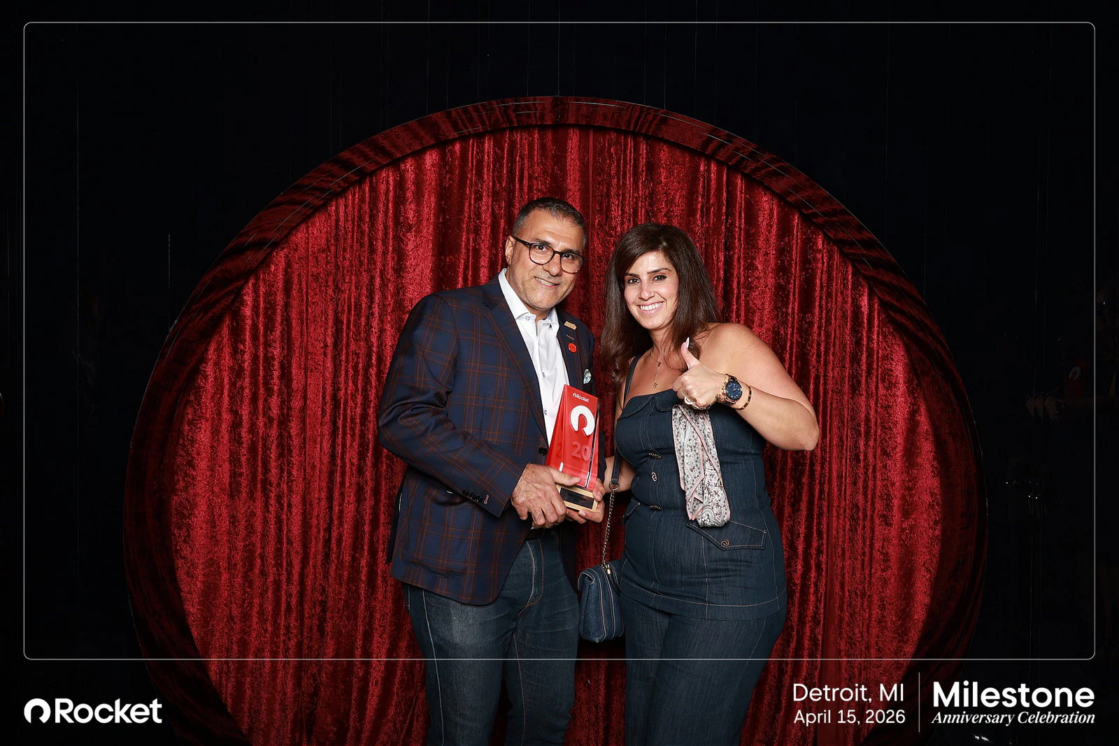 A man in a check blazer holding a red award plaque and a woman in a denim jumpsuit giving a thumbs up in front of a spotlit red velvet curtain at the Milestone Anniversary Celebration