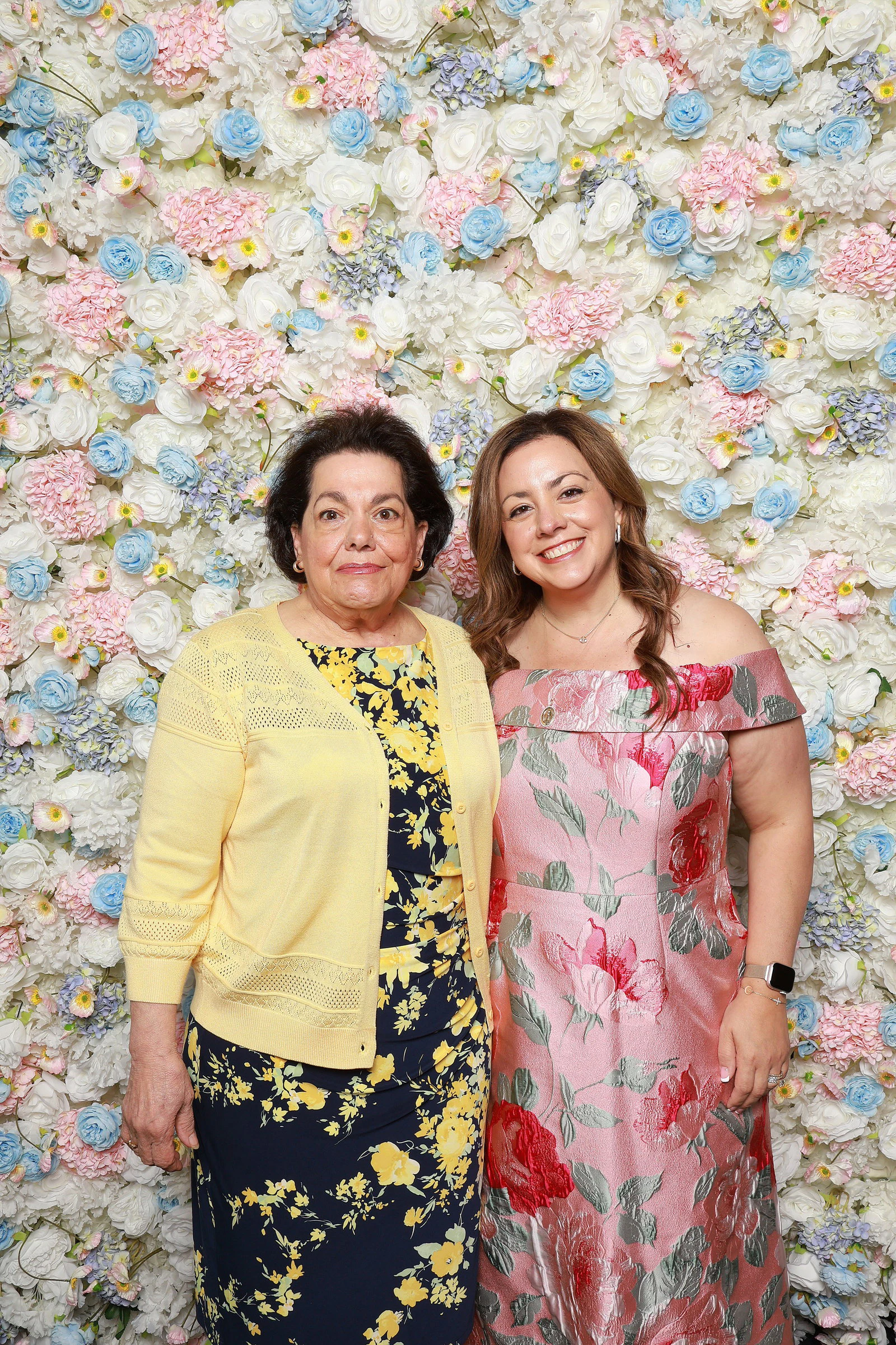 Two women cheek to cheek, one in a yellow cardigan and one in a pink floral dress, in front of the white, pink and baby blue flower wall at Mother’s Day at DAC