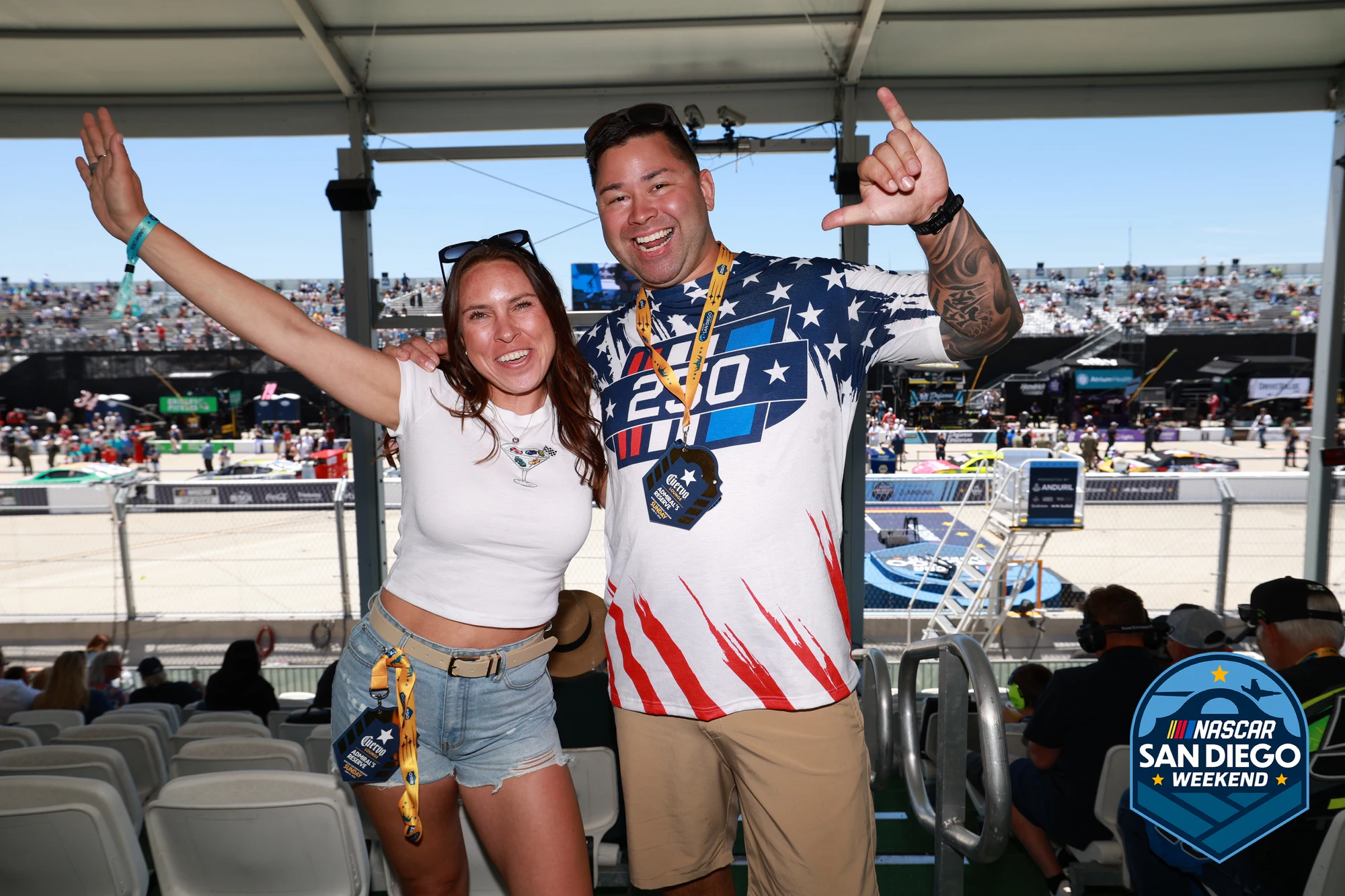Couple in flag gear, arms wide, track behind them at NASCAR San Diego