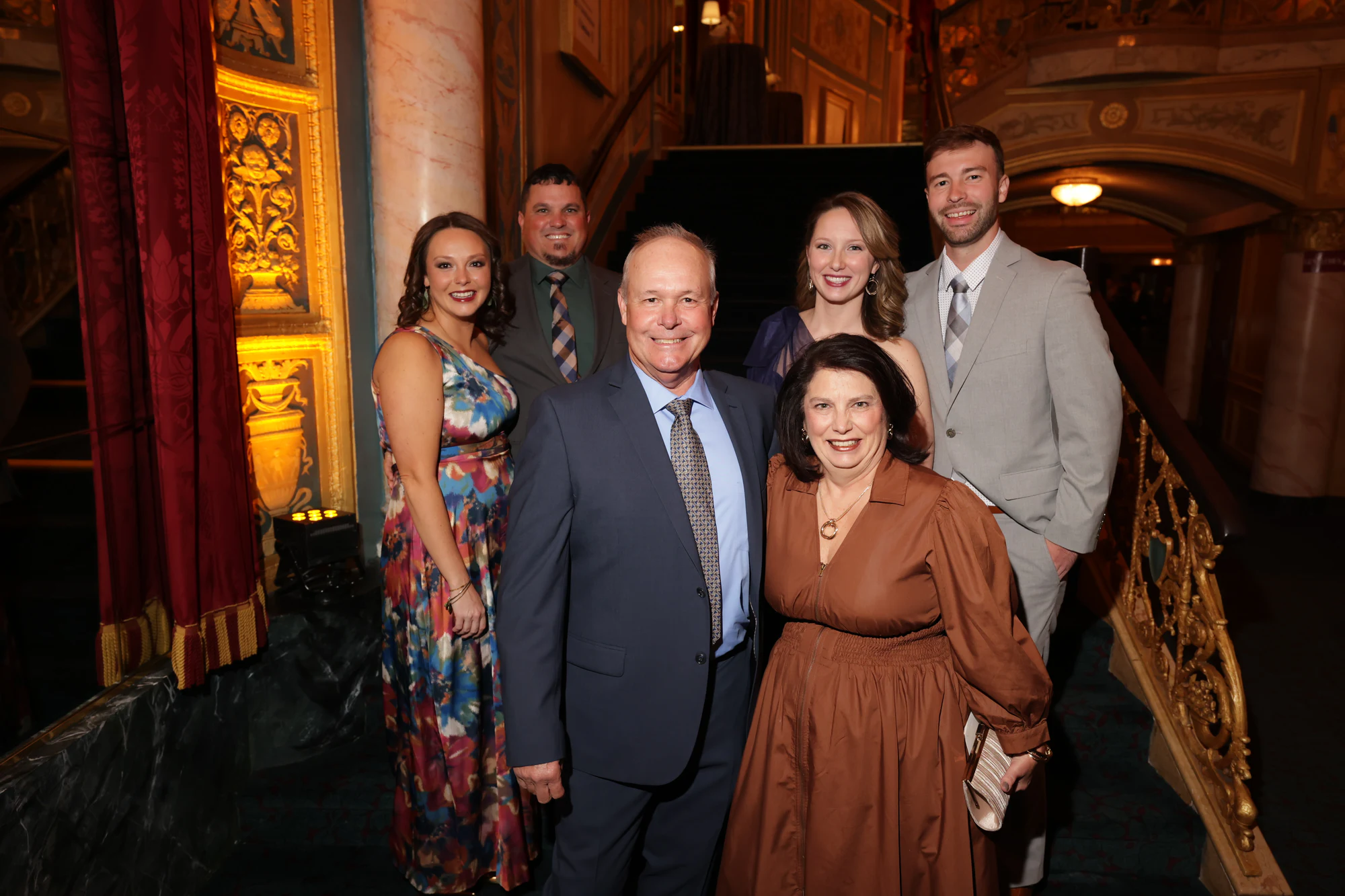 Six guests in suits and dresses grouped by the staircase at the surprise engagement party at the Detroit Opera House