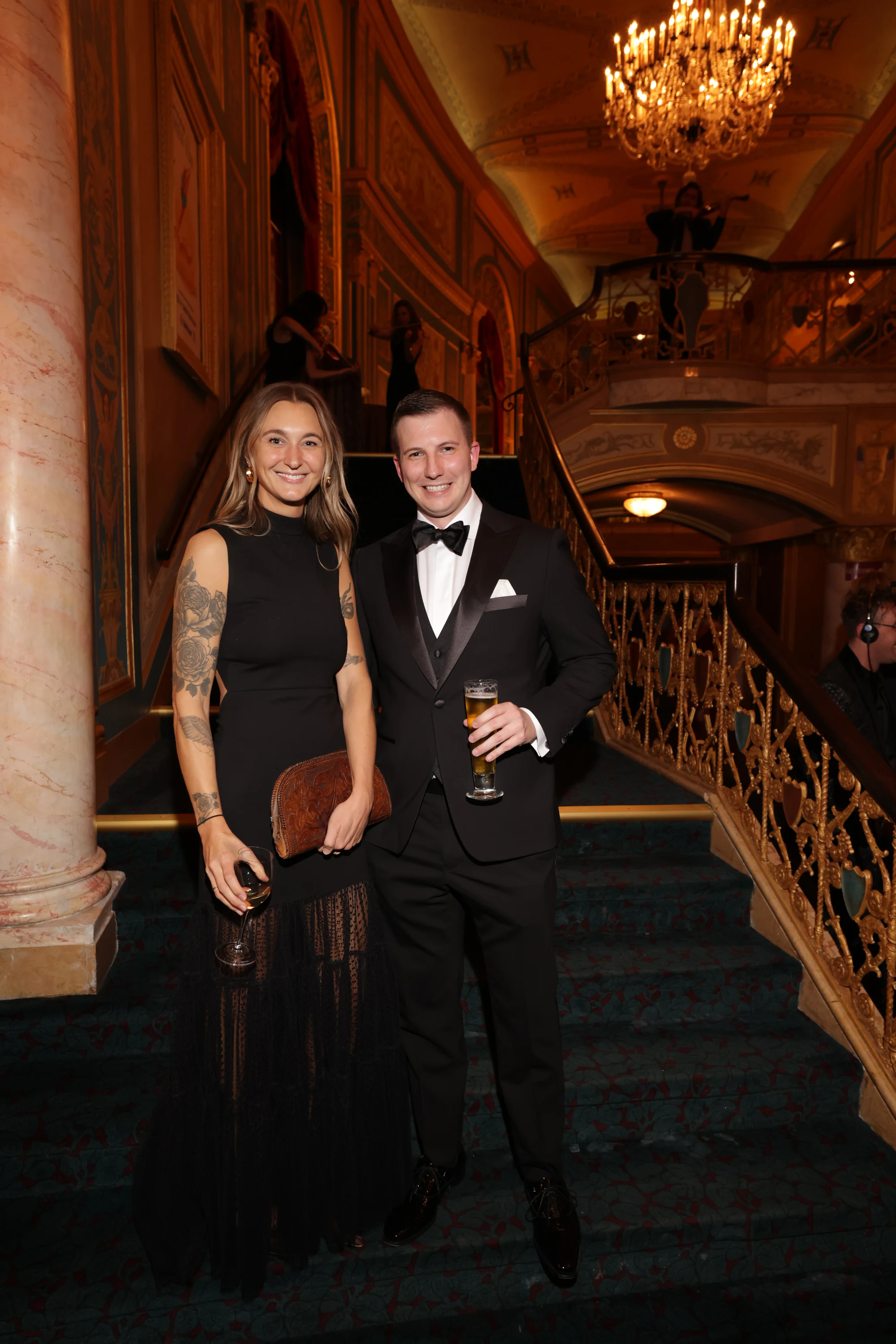 A couple on the staircase, him in a tuxedo holding a beer at the surprise engagement party at the Detroit Opera House