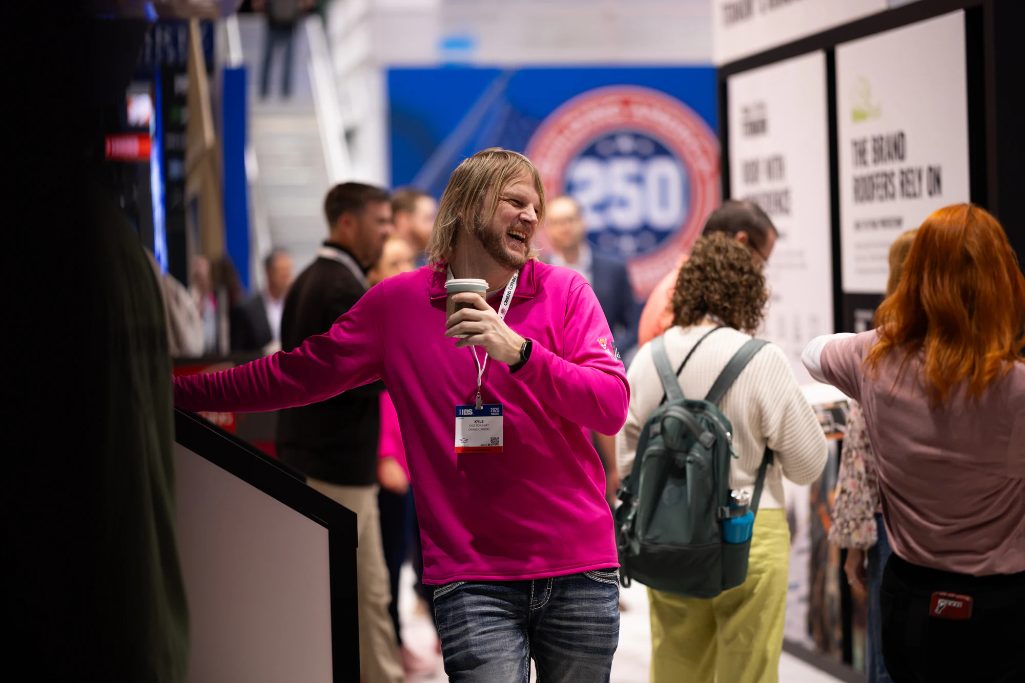 Man in the pink pullover walking the aisle with coffee, smiling at Owens Corning at IBS 2026