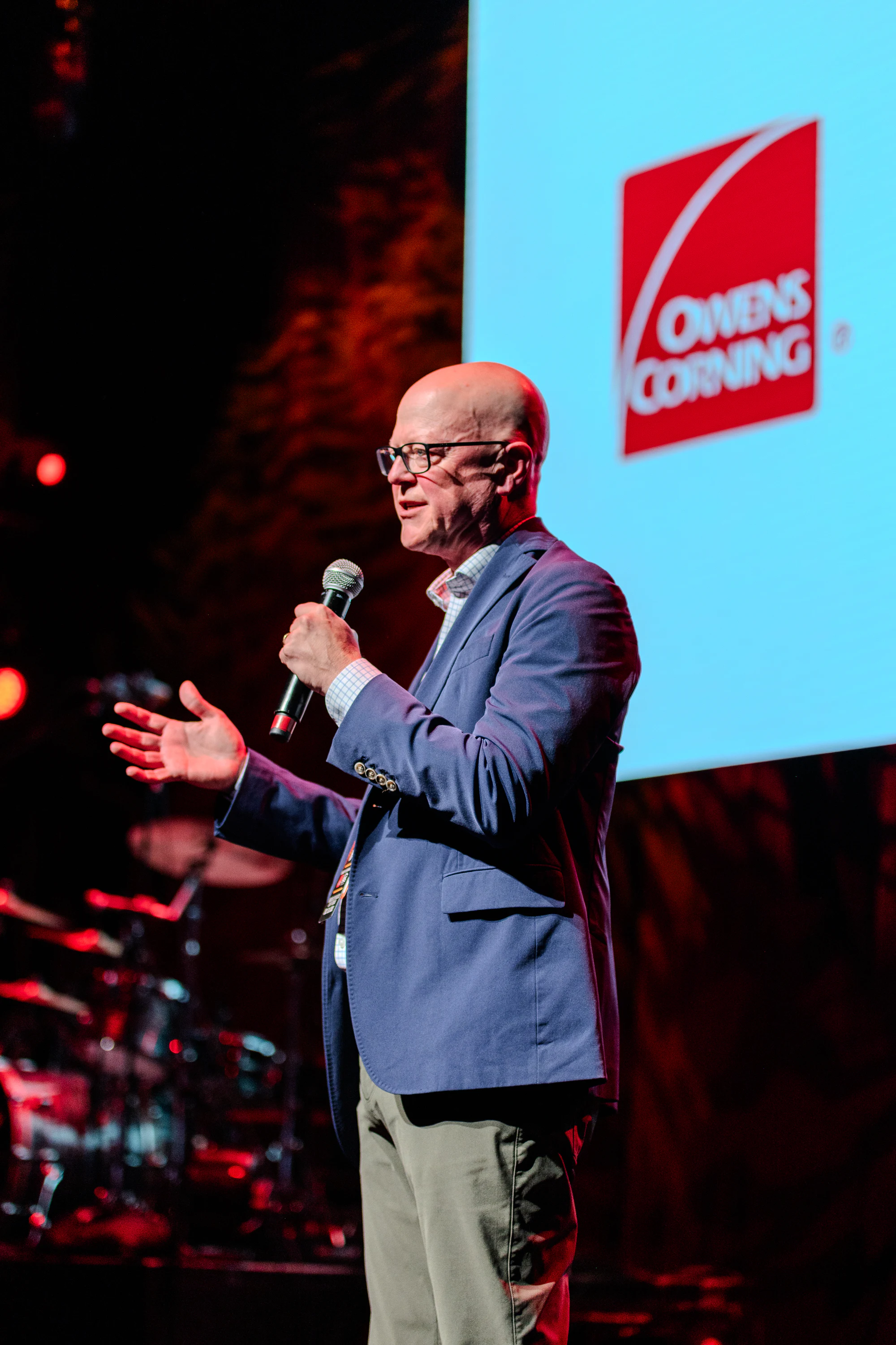Speaker with a mic on stage, a big Owens Corning logo on the screen beside him at Owens Corning at the 2023 Las Vegas Bowl
