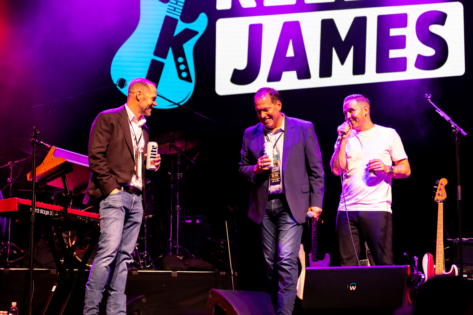 Three men sharing a laugh on stage under the Kelley James sign, one singing into the mic at Owens Corning at the 2023 Las Vegas Bowl