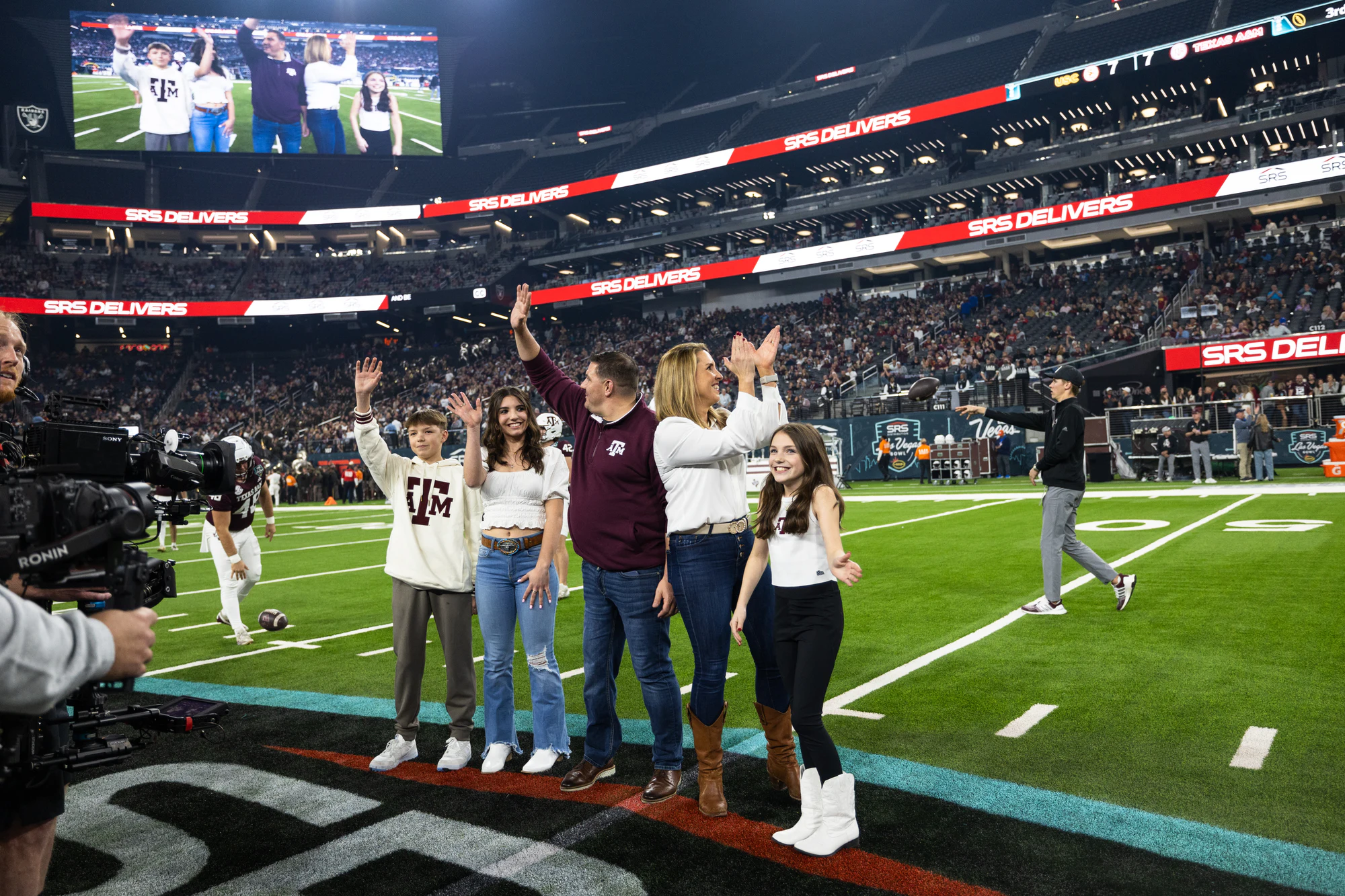 A family waves to the crowd from the field at halftime, SRS DELIVERS boards and a camera crew around them at Owens Corning at the 2024 Las Vegas Bowl