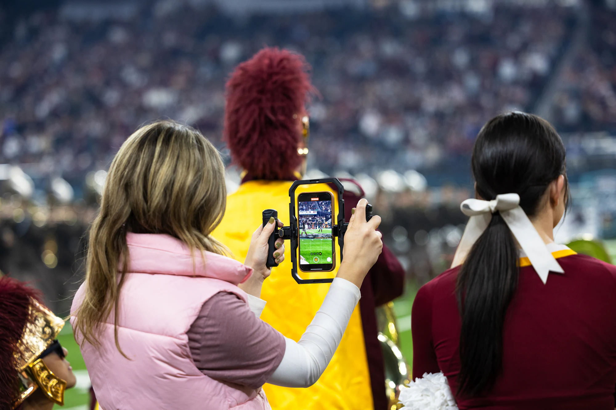 A crew member in a pink vest films the band on the sideline with a phone rig at Owens Corning at the 2024 Las Vegas Bowl
