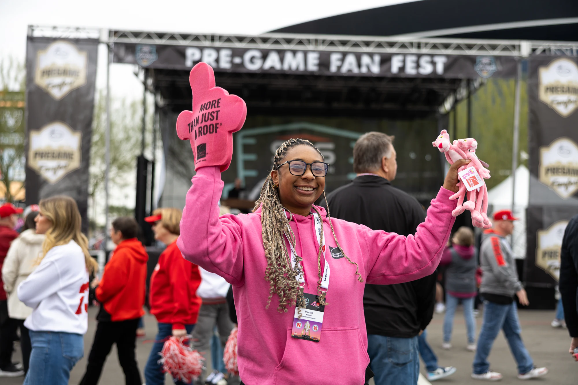 Woman in a pink hoodie holding up a pink foam finger and a Pink Panther toy in front of the Pre-Game Fan Fest stage at Owens Corning at the 2025 Las Vegas Bowl