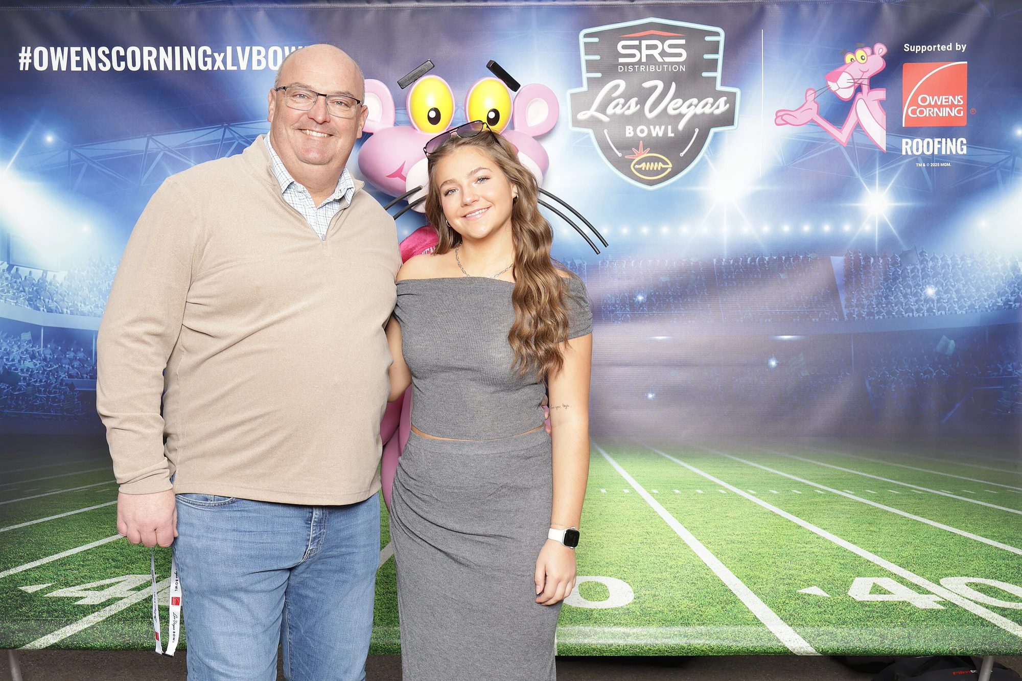 A man in a tan sweater and a woman in a gray knit set at the Fan Fest booth, Panther statue behind at Owens Corning at the 2025 Las Vegas Bowl