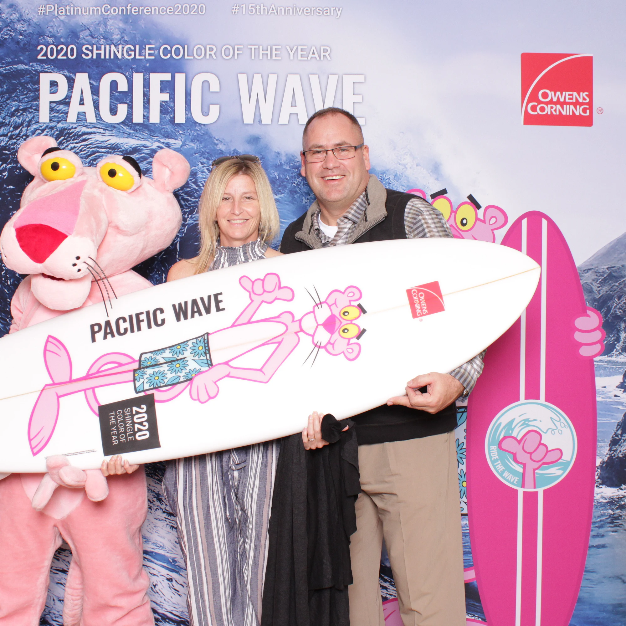 A blonde woman and a man in glasses holding the Pacific Wave surfboard beside the Pink Panther at Owens Corning Platinum Conference