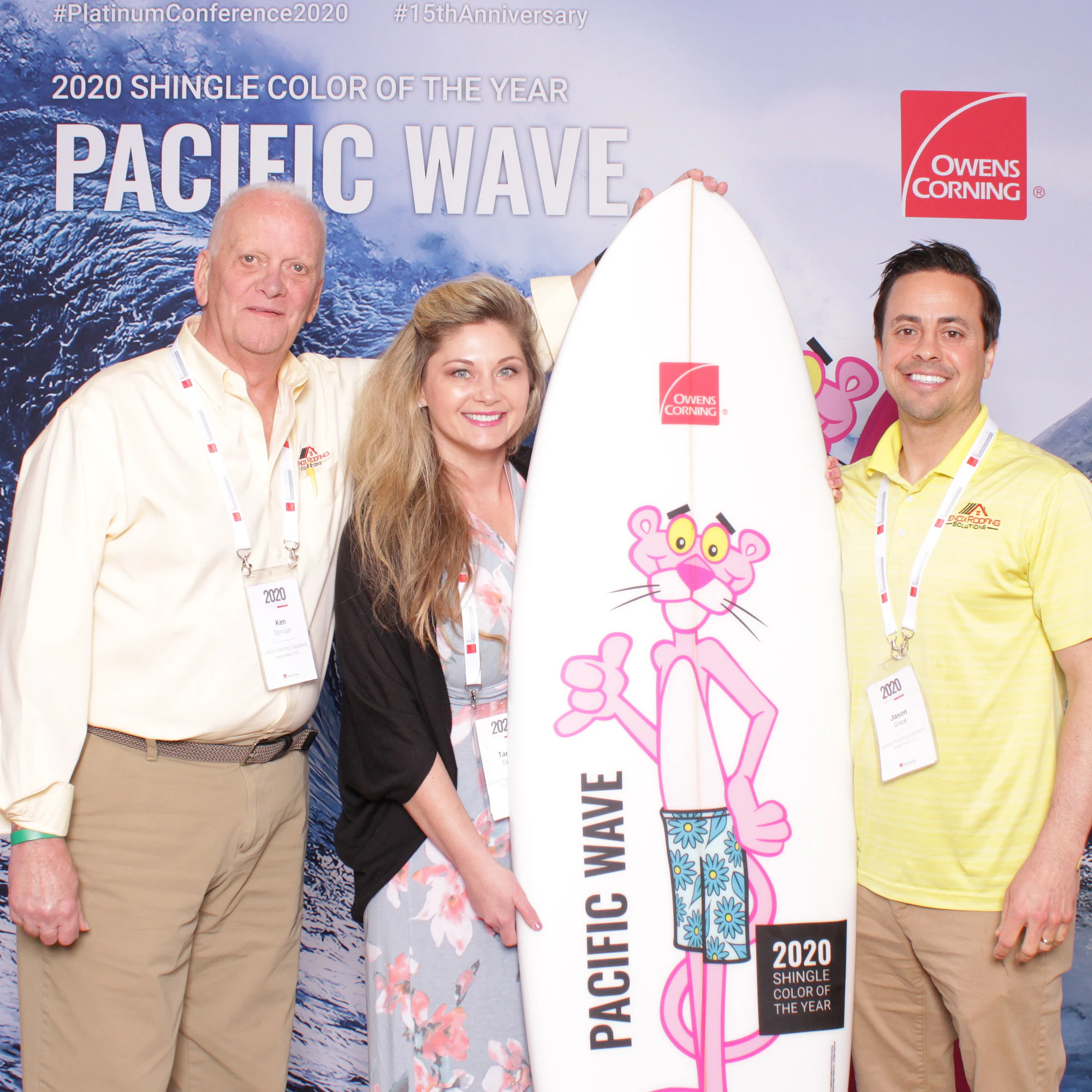 An older man in a cream shirt, a woman and a man in a yellow polo around a Pacific Wave surfboard at Owens Corning Platinum Conference