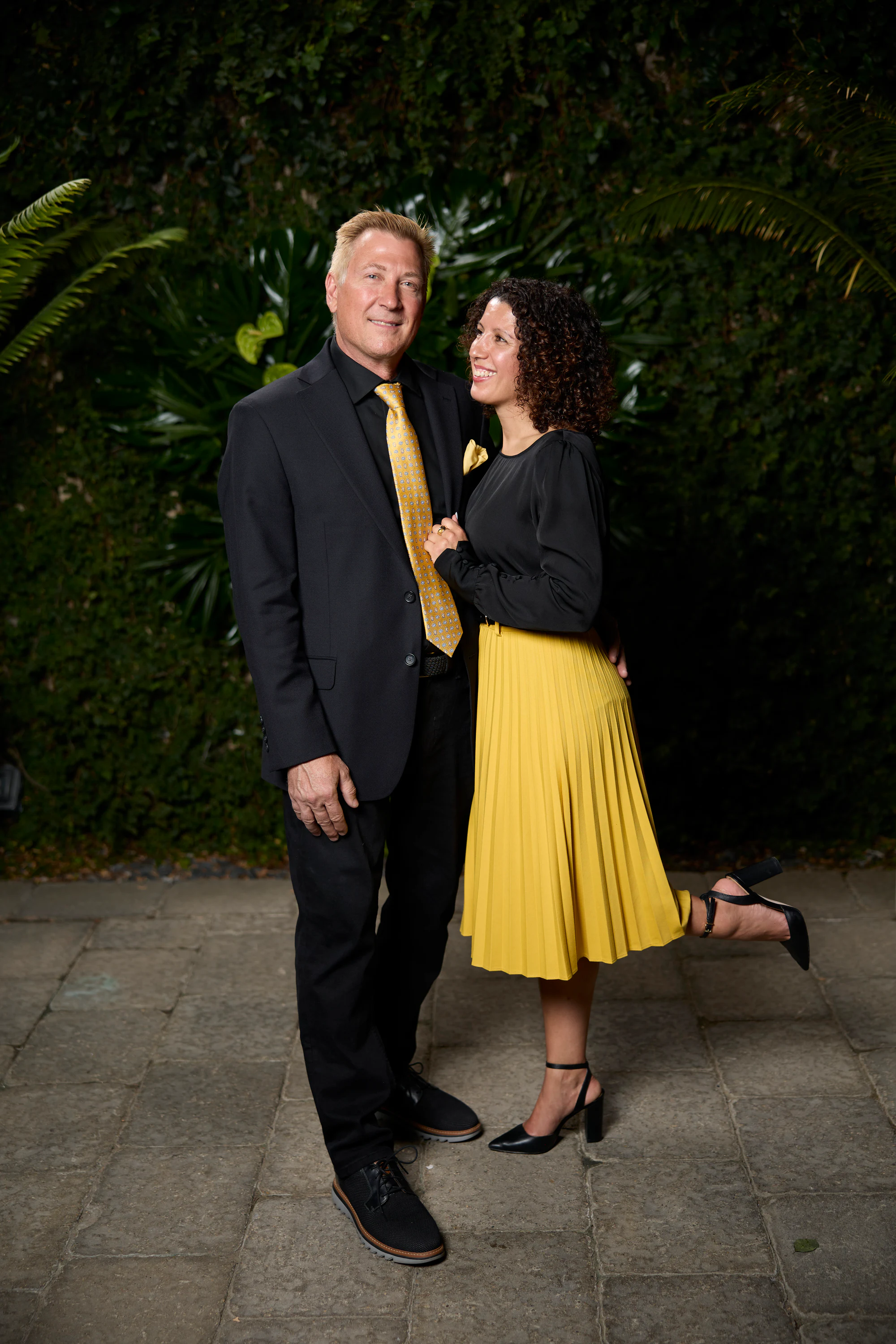 A couple, him in a black suit and yellow tie, her in a yellow skirt kicking up a heel at the Planterra Open House