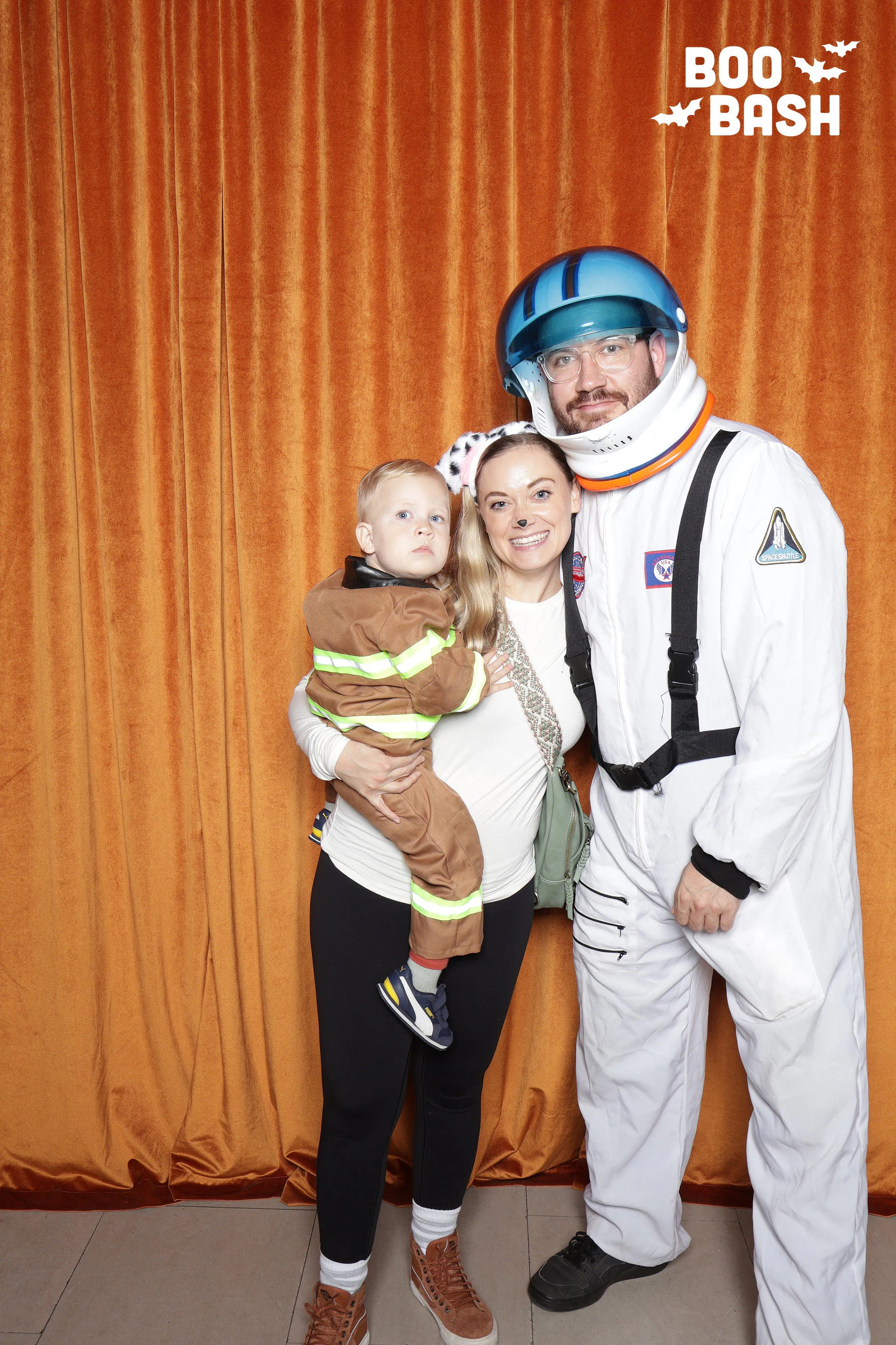 An astronaut with his helmet on beside a woman in spotted dog ears holding a toddler dressed as a firefighter at Rocket Boo Bash