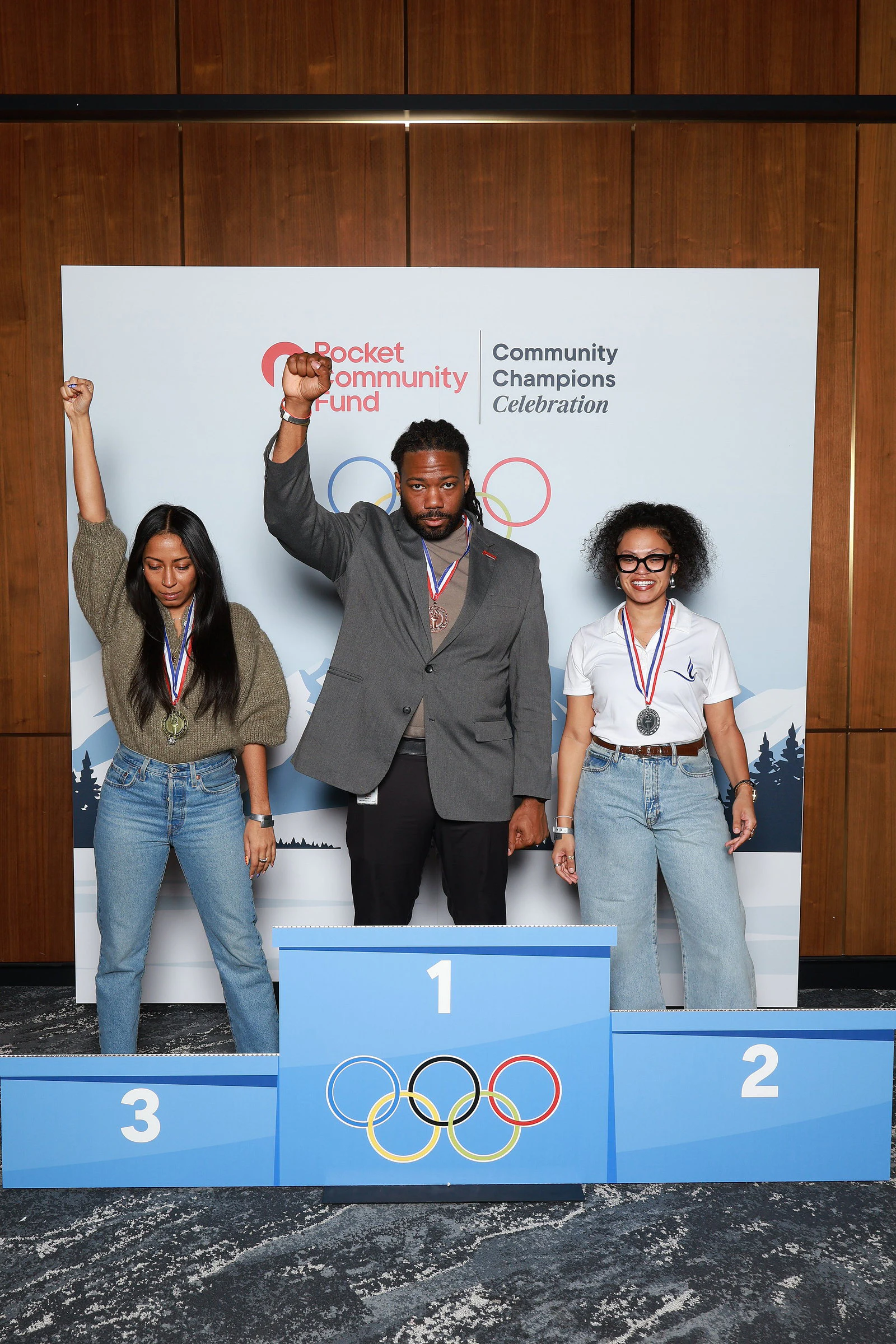 Three guests wearing medals, two with a fist raised, behind a blue winners’ podium in front of a snowy mountain backdrop at Rocket Community Champions