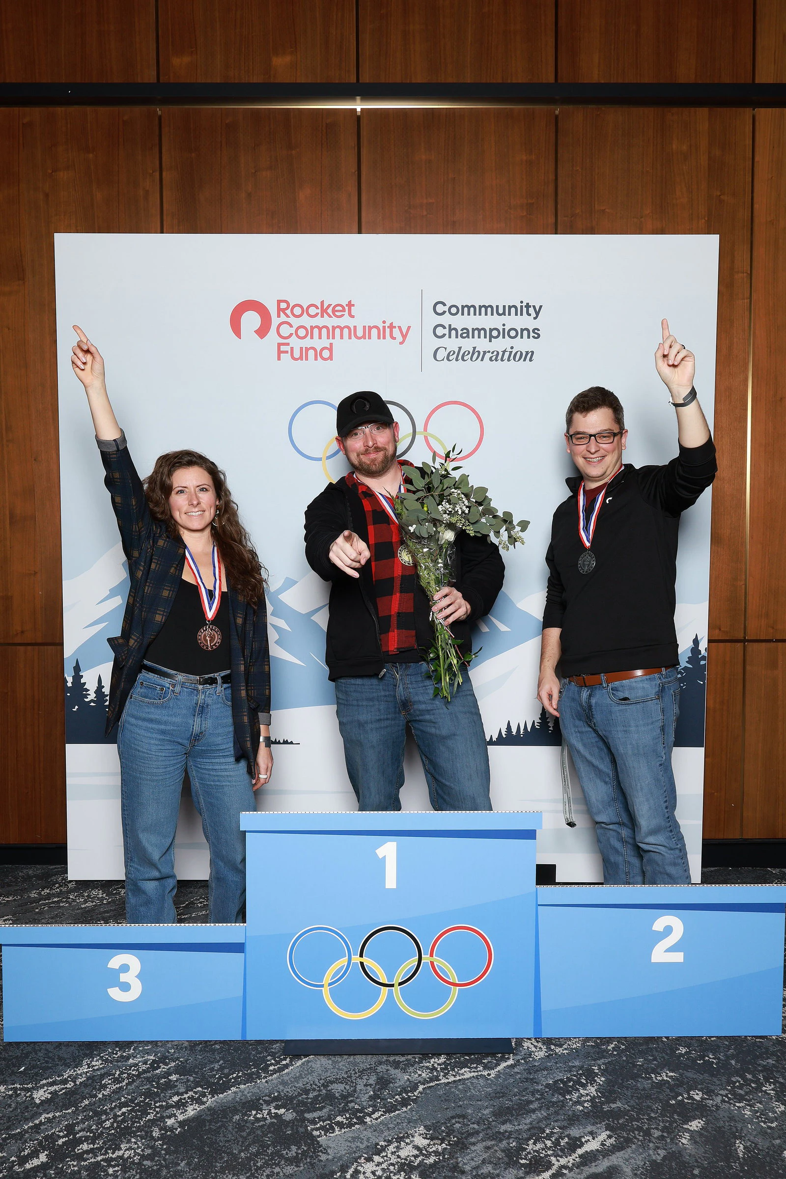 Three guests wearing medals, two pointing to the sky and one holding a bunch of greenery at Rocket Community Champions