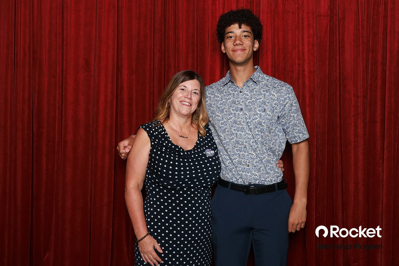 A woman in a navy polka-dot dress with her arm round a tall young man in a patterned shirt at Intern Open House with Rocket