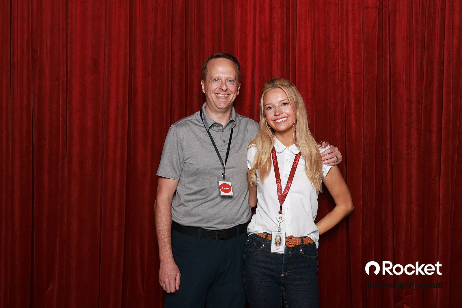 A man in a grey polo with his arm around a blonde woman in a white polo and jeans, both wearing lanyards, in front of the red velvet at the Intern Open House with Rocket