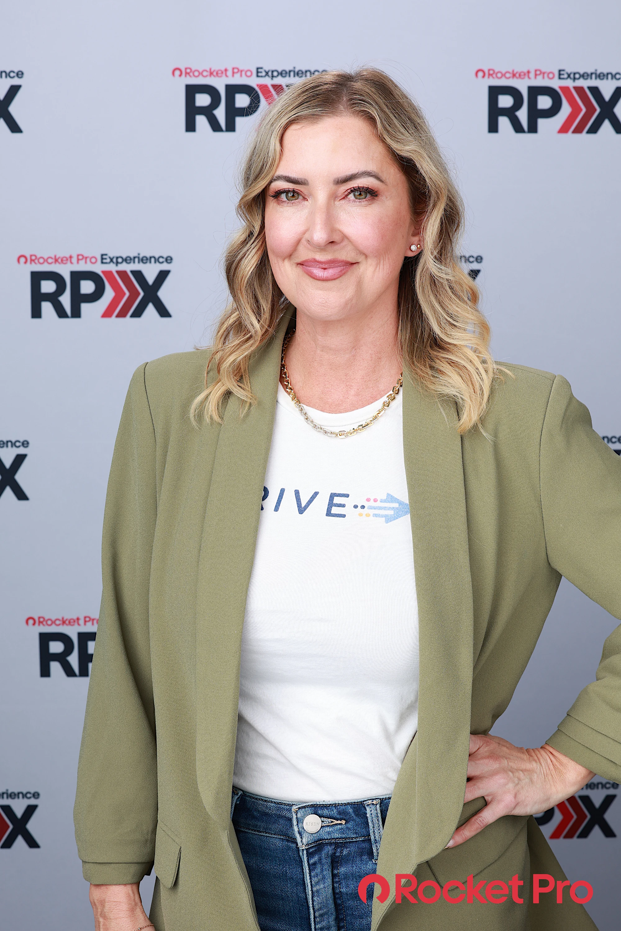 A woman in an olive blazer smiling, hand on her hip, in front of the RPX backdrop at Rocket Pro Experience