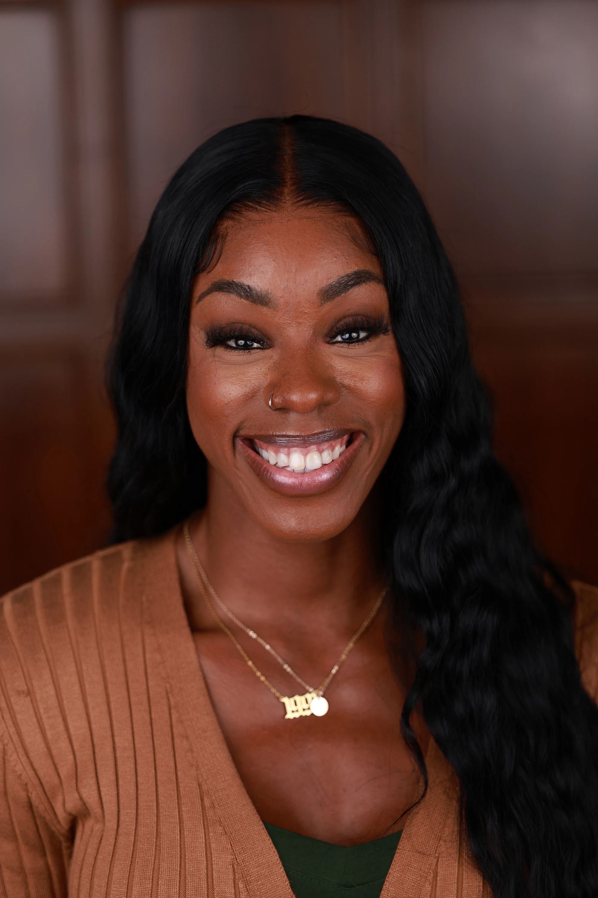 Headshot of a woman with long dark hair and a tan cardigan, smiling wide, against a wood-paneled wall at Remote Welcome Reception