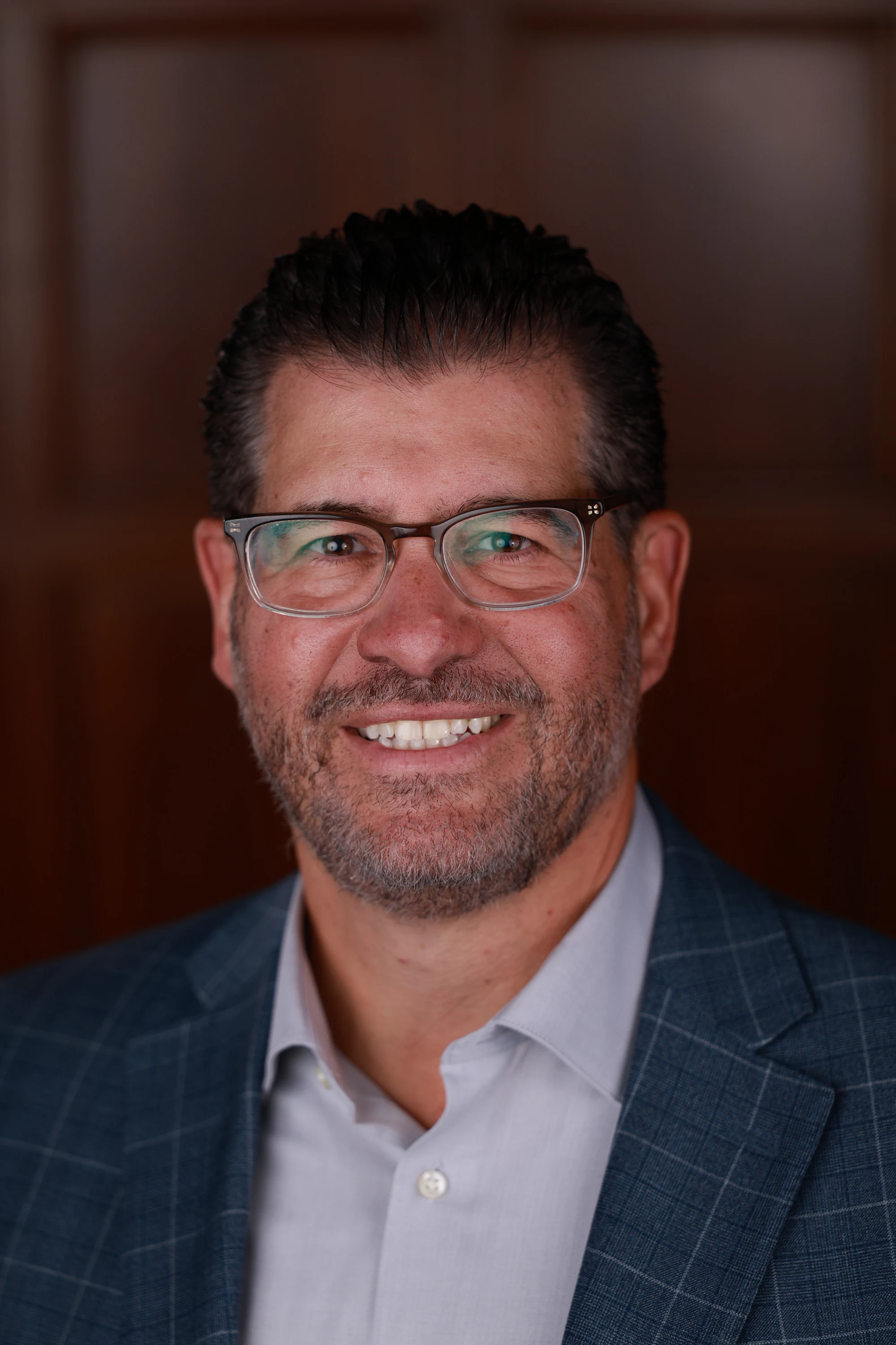 Headshot of a man in glasses and a blue check blazer against a wood-paneled wall at Remote Welcome Reception