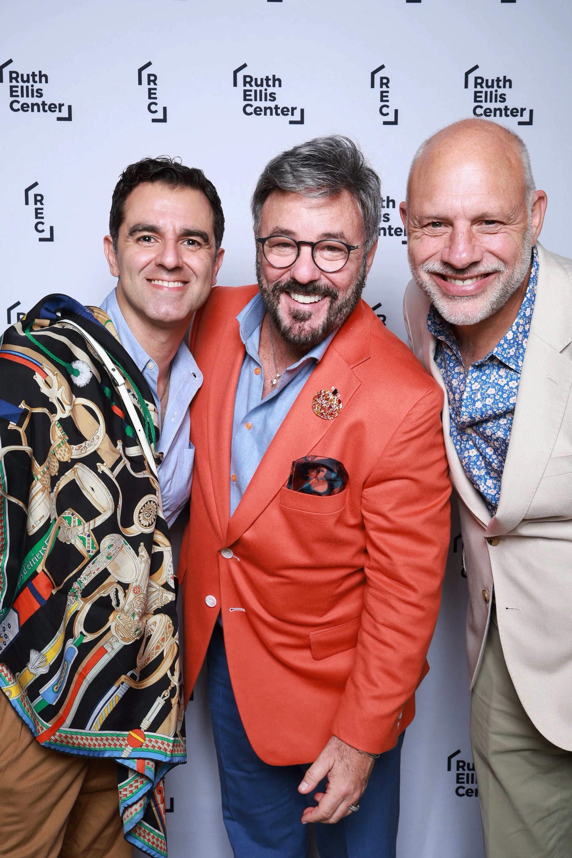 Three men smiling shoulder to shoulder, one in an orange blazer and one with a printed scarf over his shoulder at Ruth Ellis Center Annual Gala