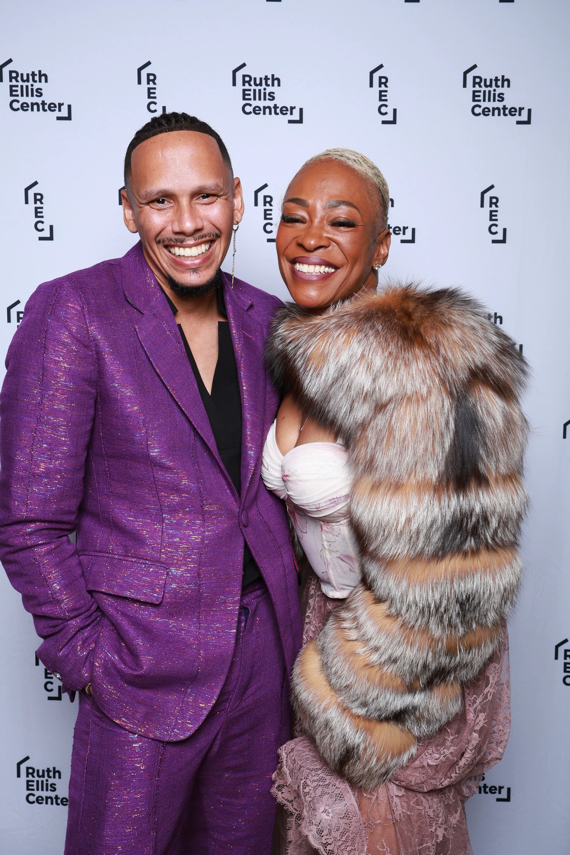 A man in a purple suit and a woman in a fur coat smiling in front of the Ruth Ellis Center backdrop at Ruth Ellis Center Annual Gala