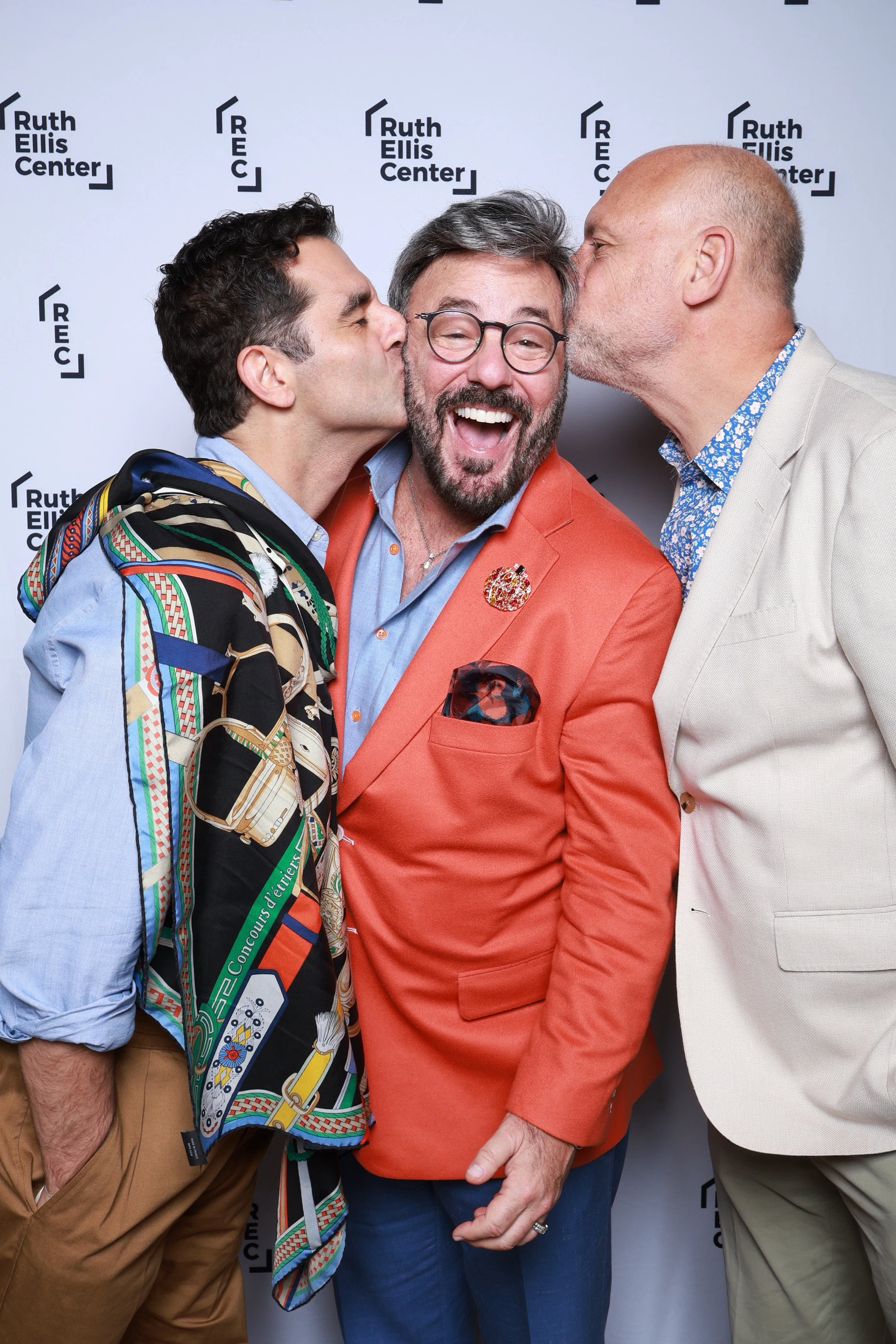 Two men kissing the cheeks of a laughing man in an orange blazer at Ruth Ellis Center Annual Gala