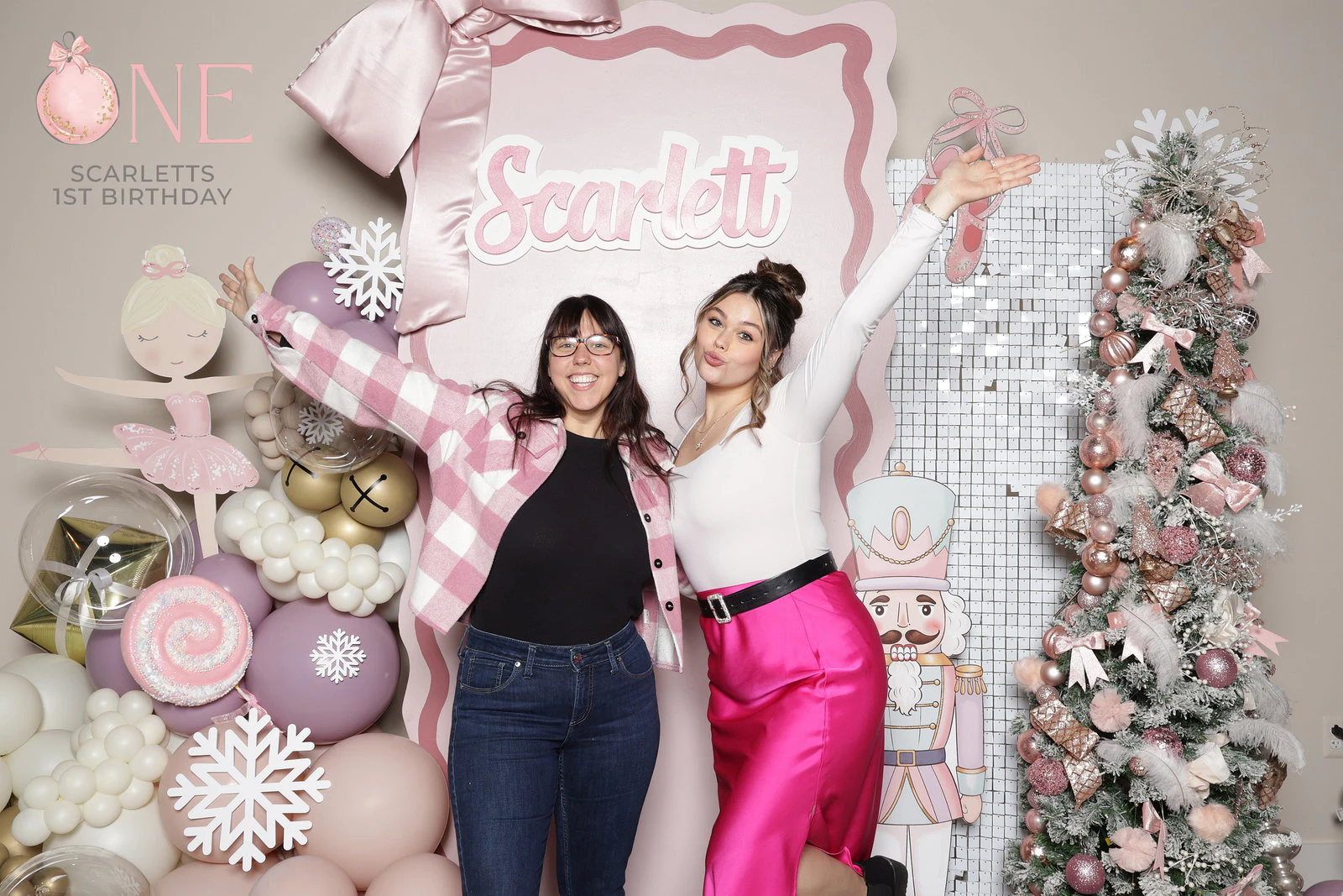 Two women with their arms flung out, one in a pink check jacket and one in a white top and bright pink skirt, in front of a pink ballerina and nutcracker birthday set at a private party