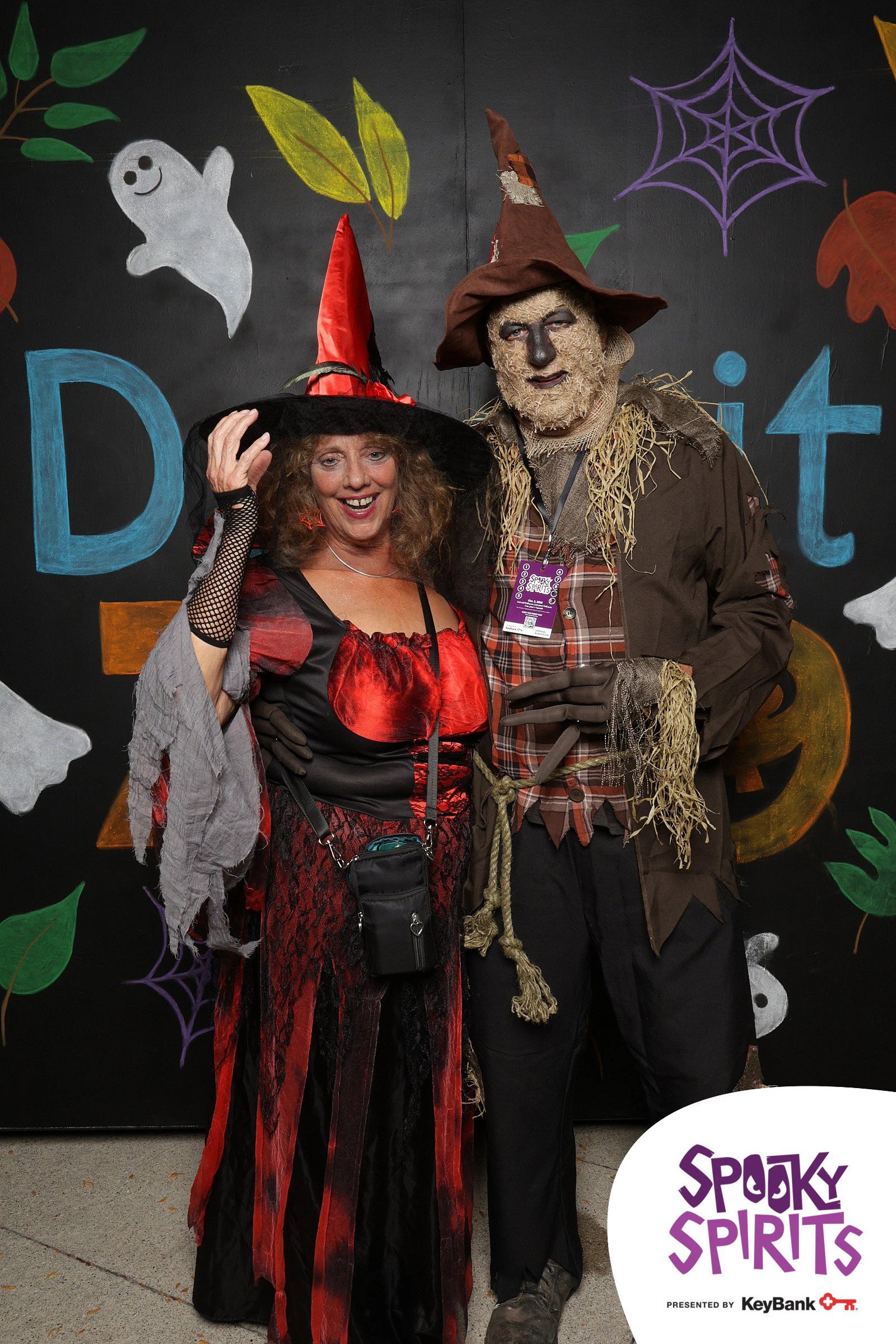A witch in a red hat and a masked scarecrow in front of a black wall painted with ghosts, cobwebs, leaves and pumpkins