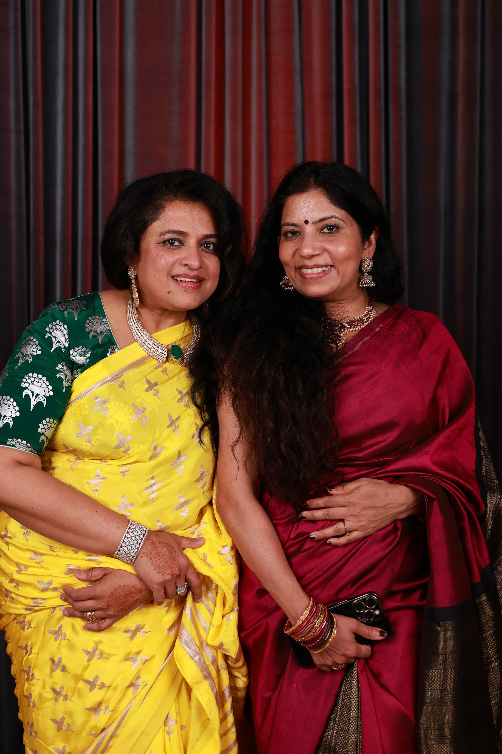 Two women in a yellow and a red sari, arm in arm at Puja + Curtis at State Savings Bank