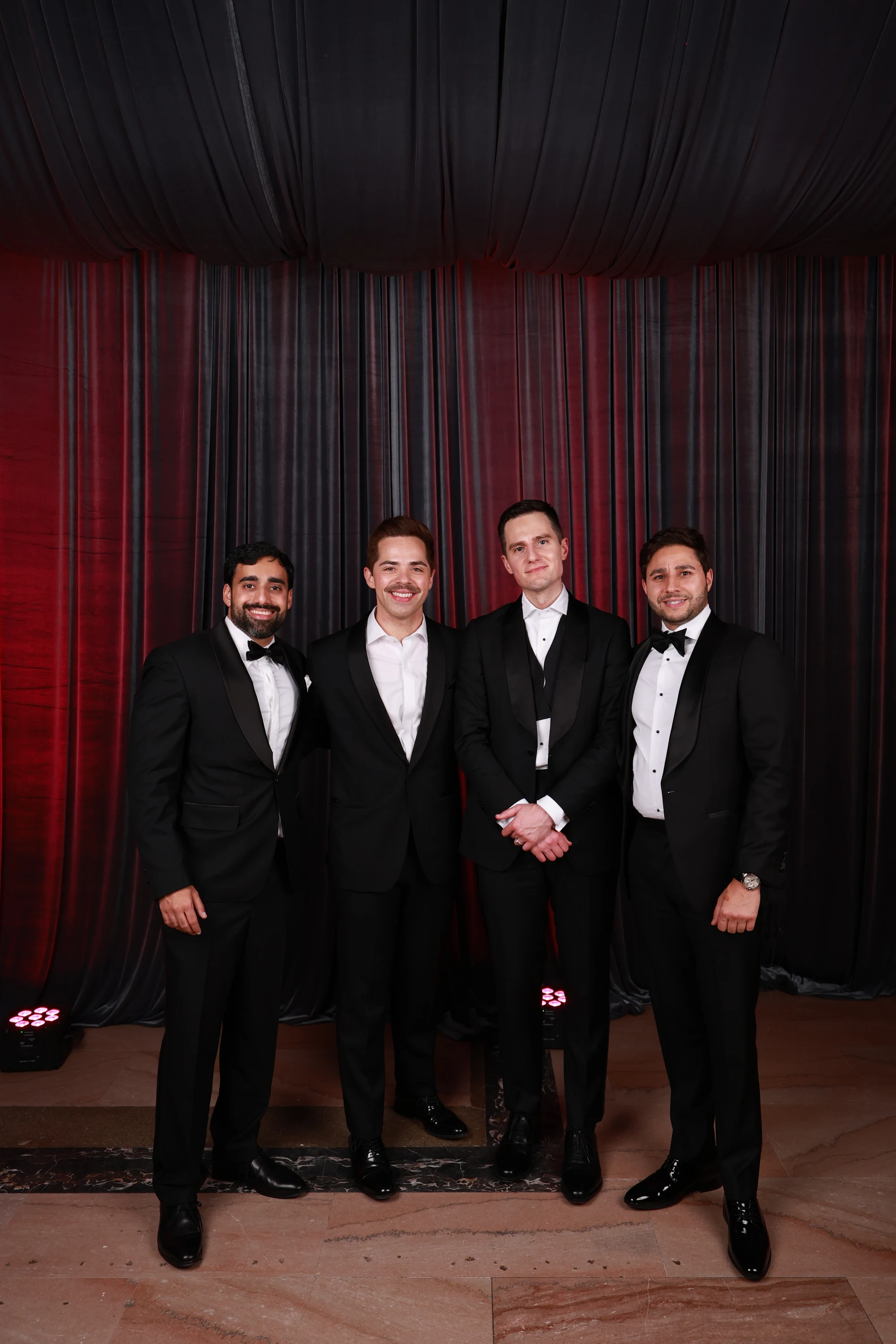 Four men in tuxes lined up, red wash behind at Puja + Curtis at State Savings Bank