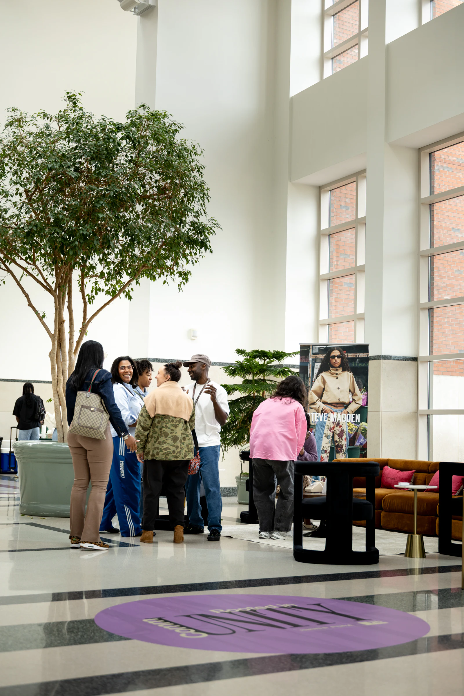 Guests talking beside the Steve Madden lounge in a tall white atrium, a purple decal on the floor in front at Black Footwear Forum